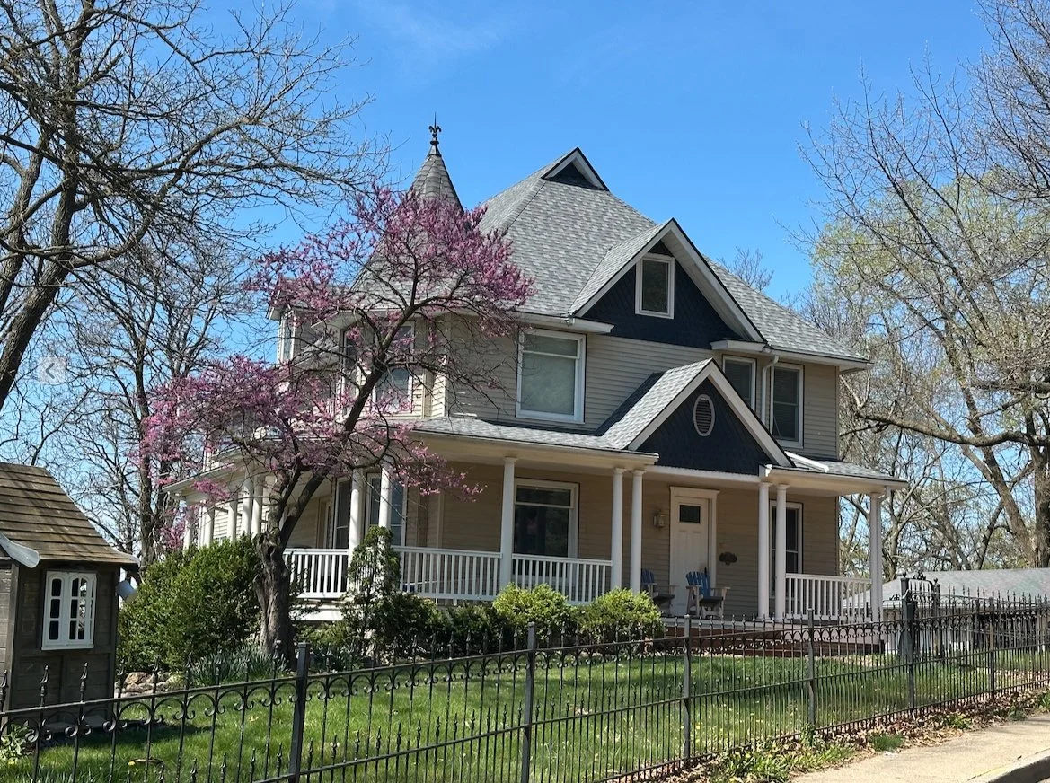 A large, Victorian-style house with a front porch, surrounded by a black metal fence and trees, one with pink blossoms, under a clear blue sky.