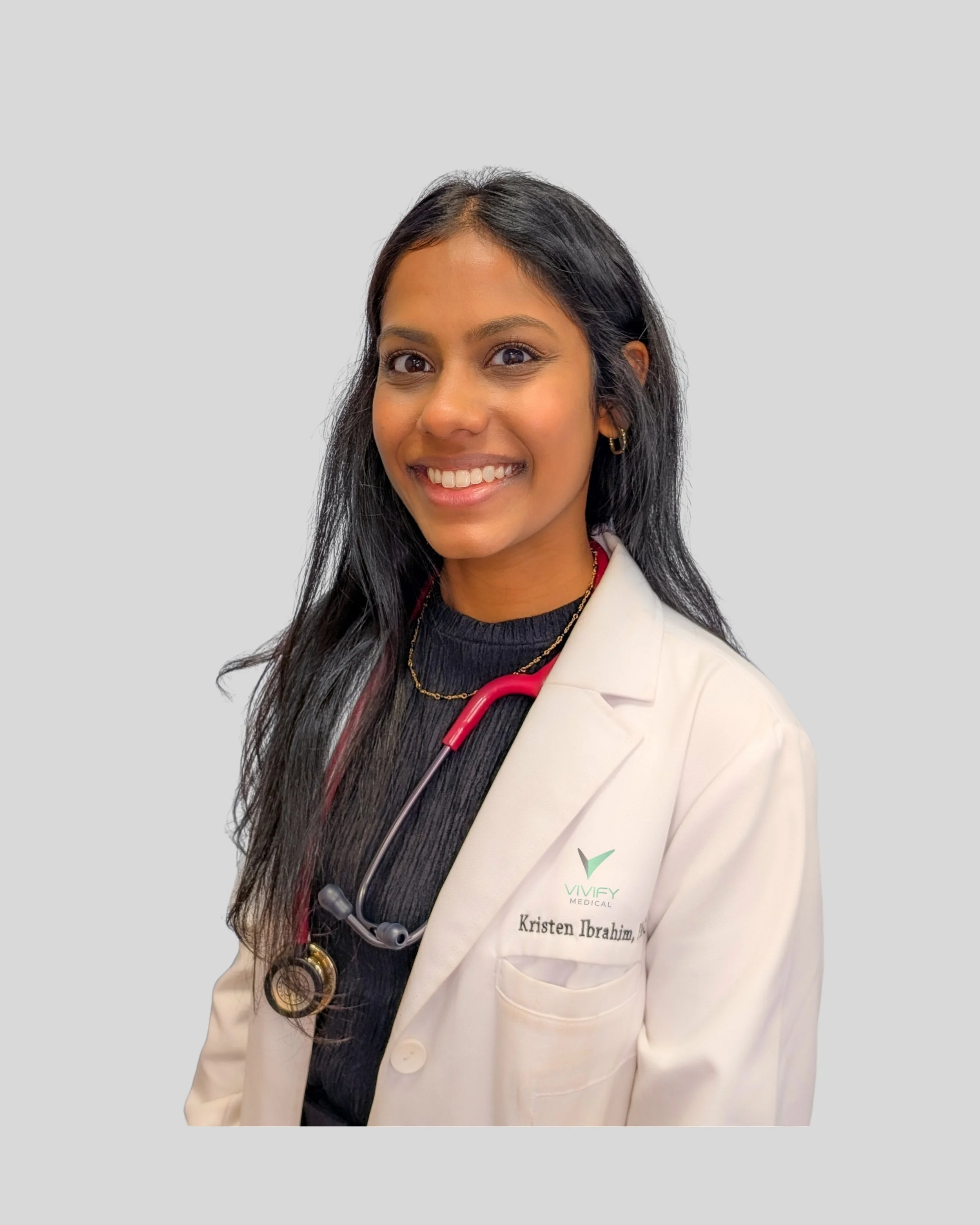 A smiling woman with long black hair wearing a medical white coat, a stethoscope, and gold jewelry, against a plain light gray background.