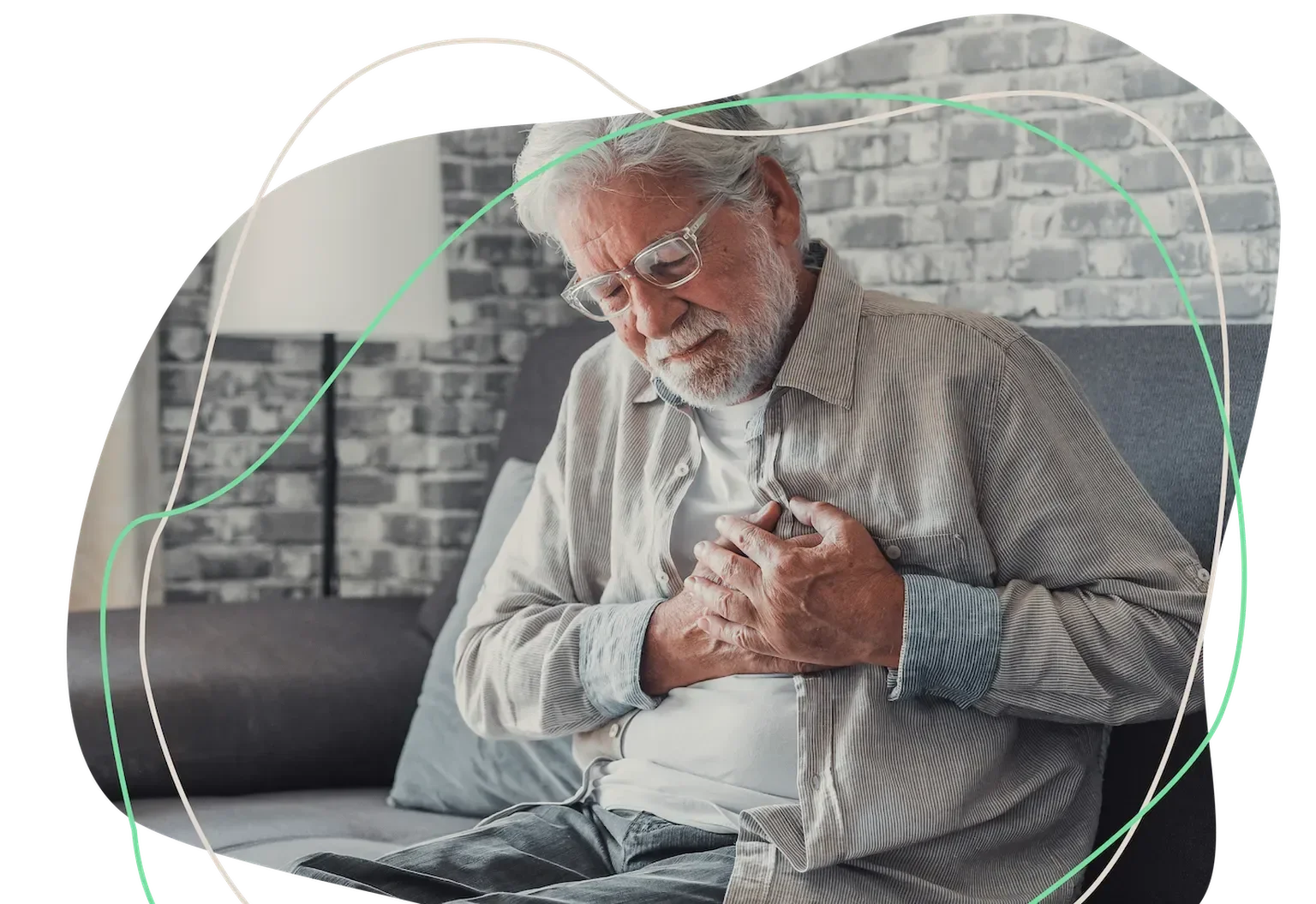 An elderly man with glasses sitting on a gray couch clutching his chest, indicating chest pain or discomfort, in a living room with brick wall background.