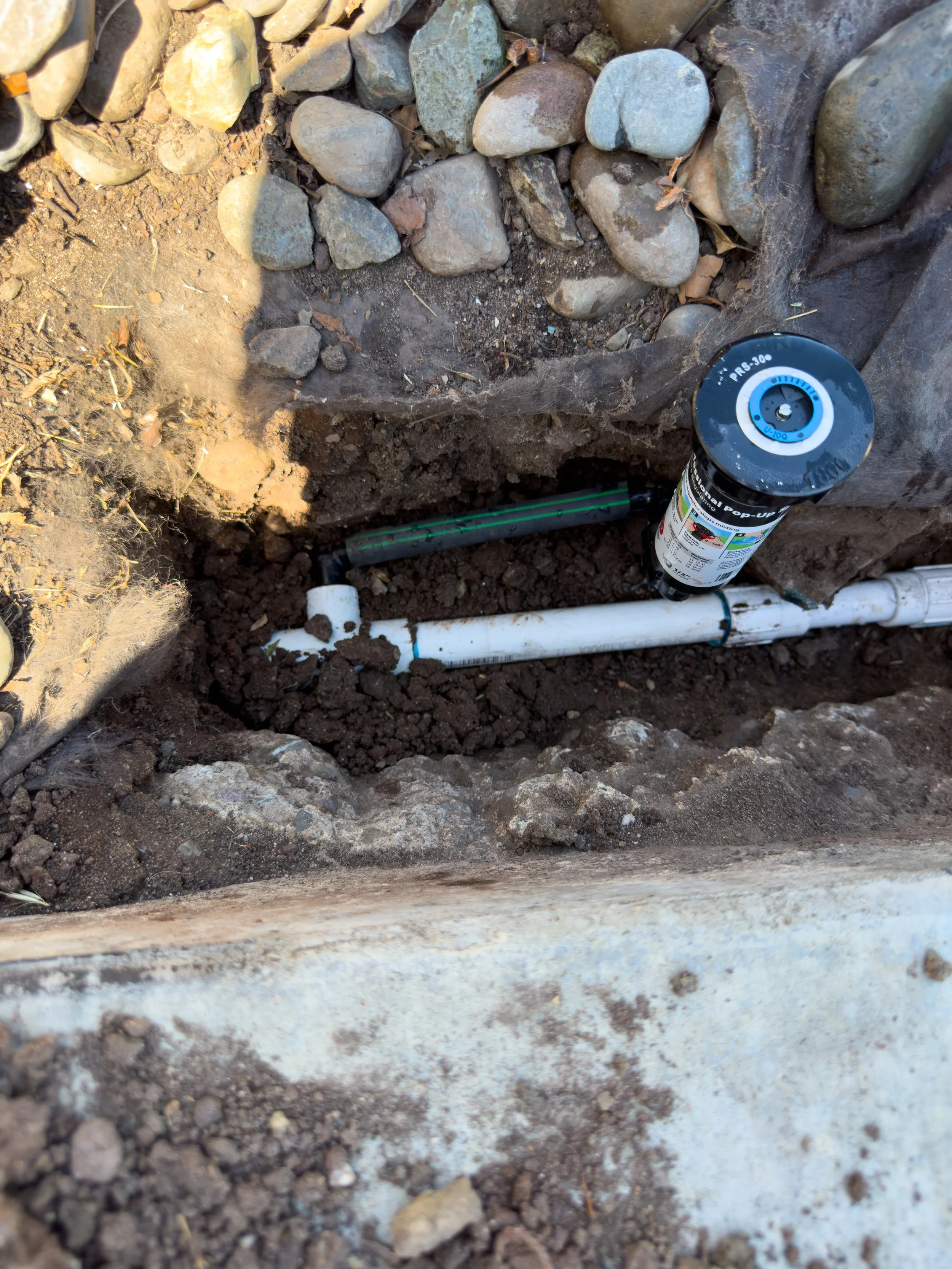 Close-up of a sprinkler repair involving white plastic pipes, a swing joint, and underground wiring in a dirt trench surrounded by rocks.