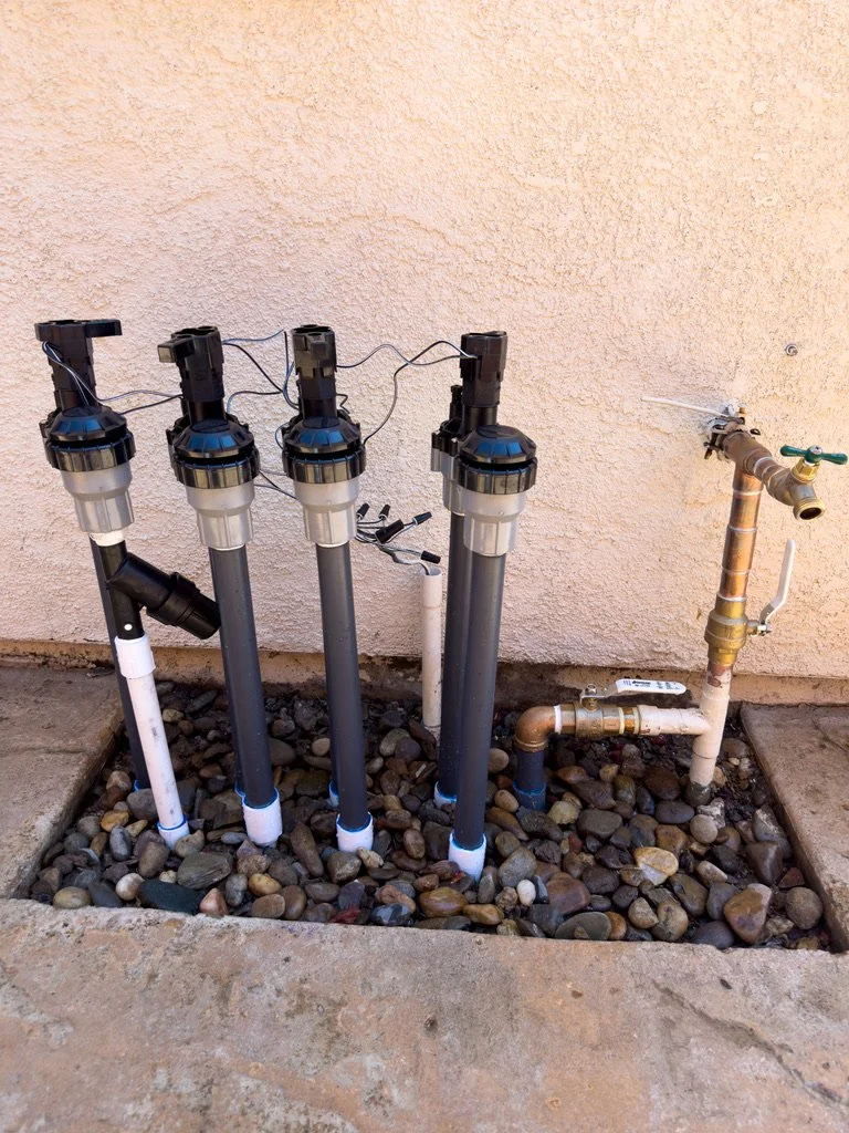 Outdoor plumbing with four black and gray rain bird valves, PVC pipes, and a brass valve assembly against a peach-colored stucco wall on a bed of rocks.
