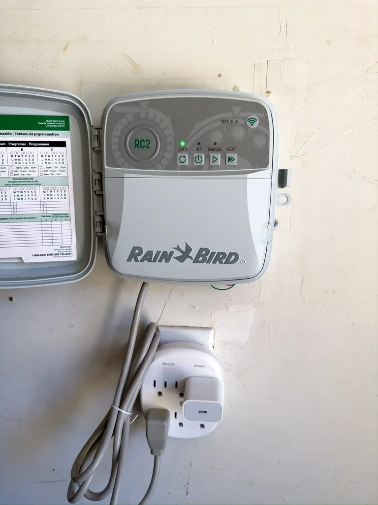 Rain Bird irrigation smart WIFI controller plugged into a power outlet, mounted on a white wall.