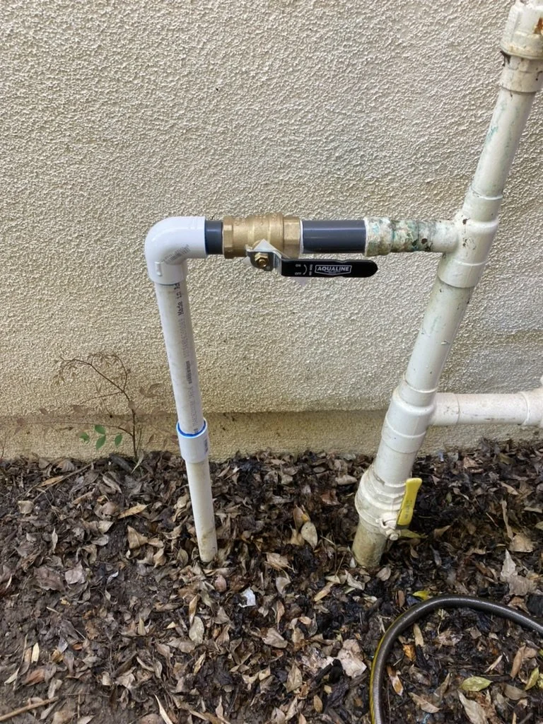 Outdoor plumbing pipes connected to a wall, with a shut-off valve labeled 'AQUALINE'. The pipes are partially surrounded by fallen leaves on the ground.