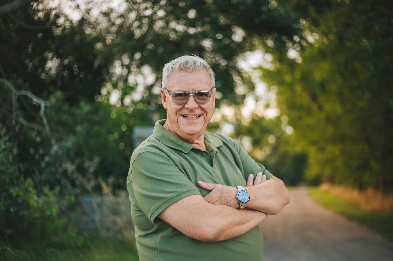 Hombre mayor sonriendo, con gafas y camiseta verde, de pie en un camino rodeado de árboles en un entorno natural.