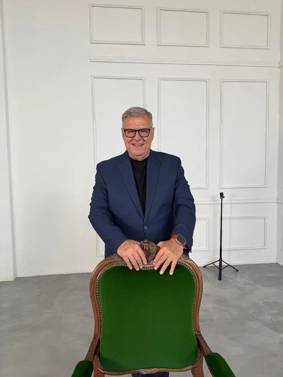 Hombre de cabello gris, gafas, ropa formal azul, sonriente, de pie detrás de una silla de madera con respaldo tapizado en verde, en una sala blanca con molduras en la pared.