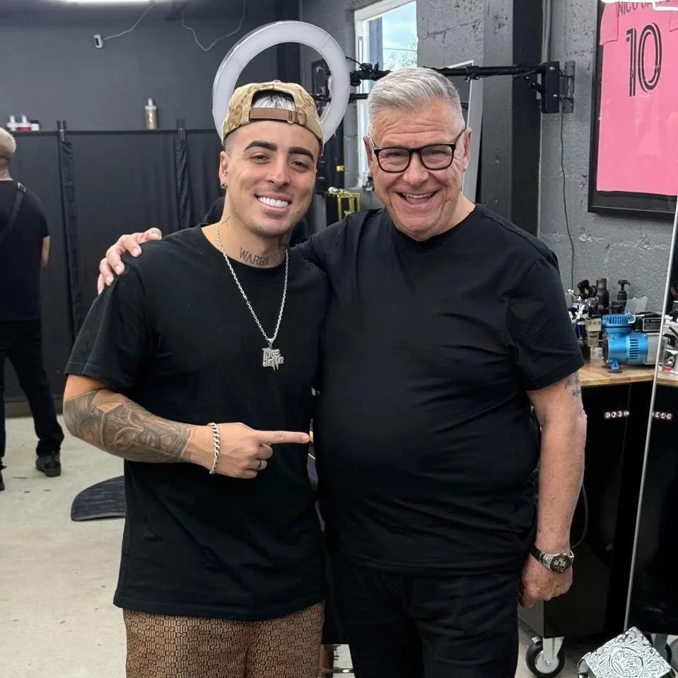 Dos hombres sonriendo y abrazándose en un estudio de tatuajes o barbería. Uno lleva gorra, collar con colgante y tatuajes, y el otro usa gafas y una camisa negra.