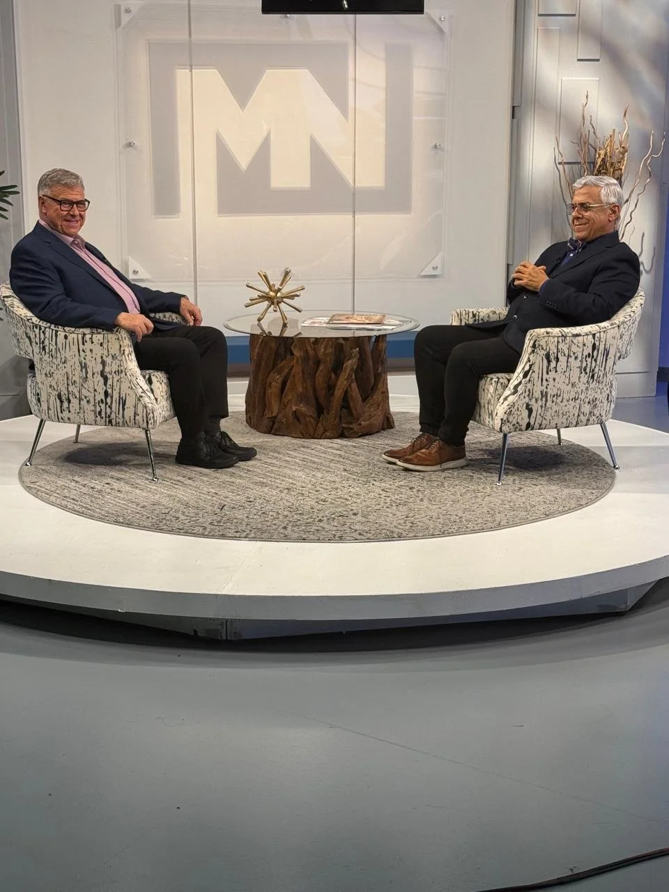 Dos hombres sentados en sillones frente a una mesa de madera en un estudio de televisión, conversando y sonriendo.
