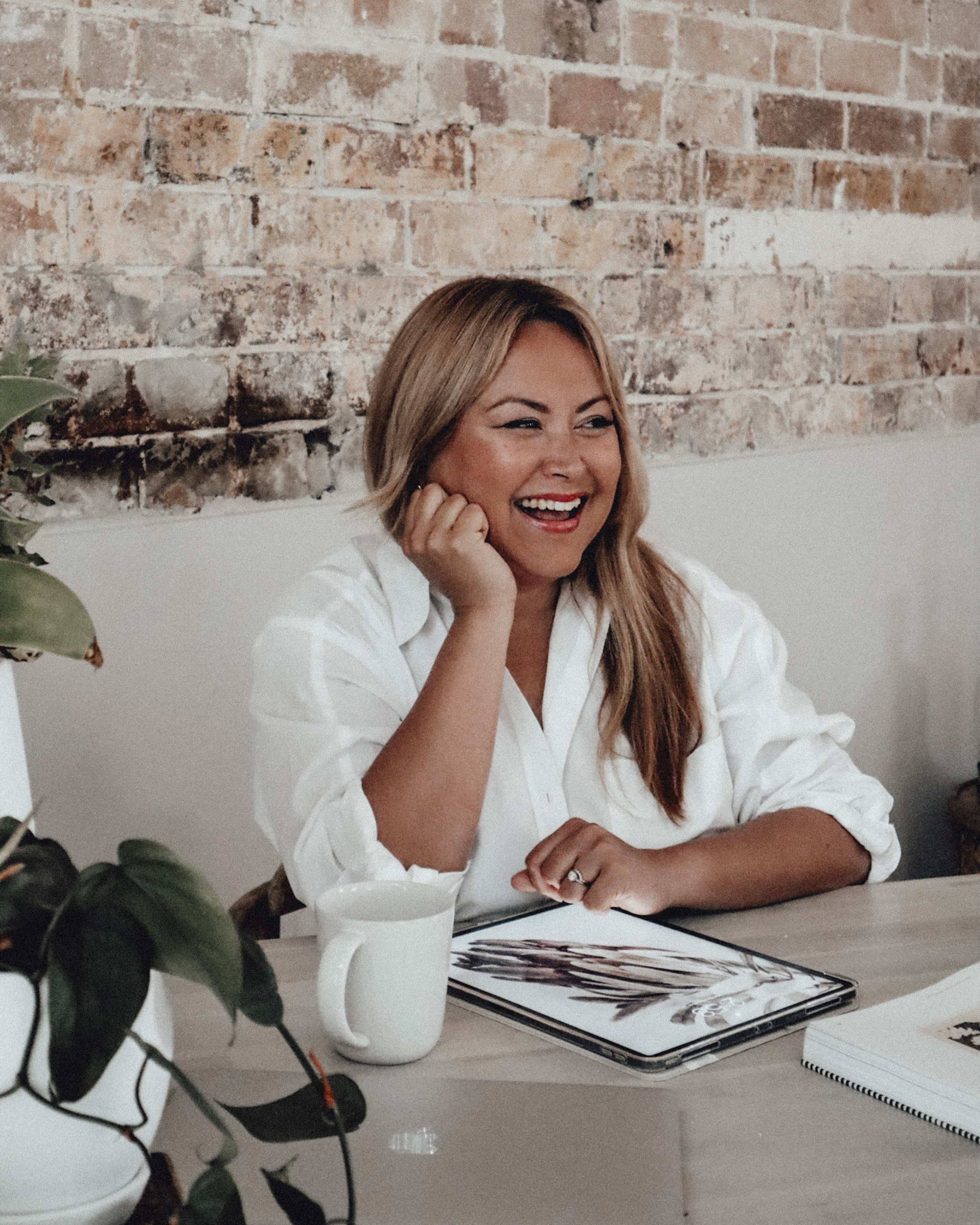 A woman laughing while sitting at a desk with a tablet, a notebook, a white mug, and a potted plant in front of a brick wall.