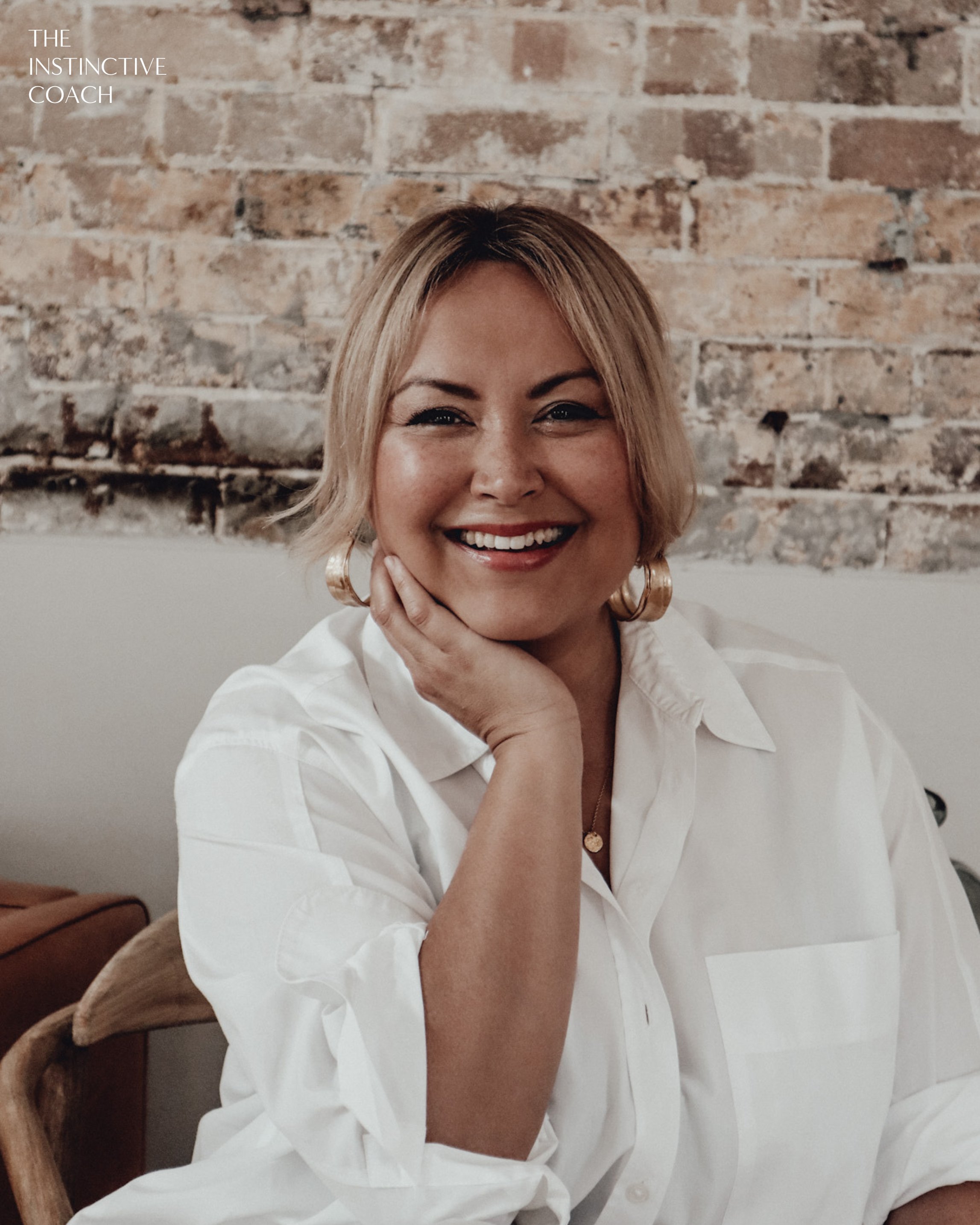 A woman with blonde hair smiling, wearing a white shirt, sitting against a brick wall background.