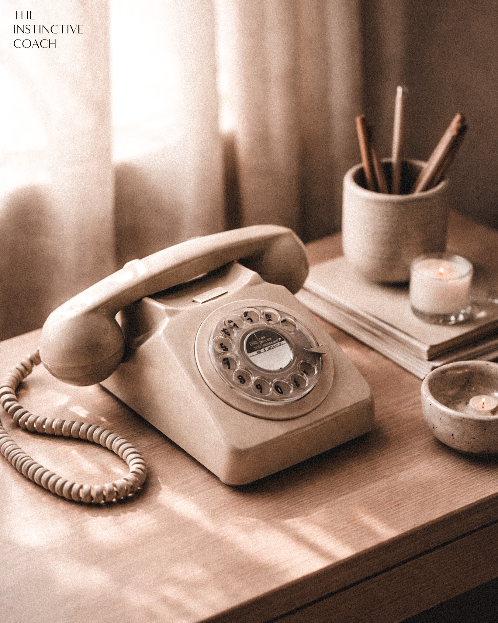 Vintage beige rotary phone on a wooden desk, with a small lit candle, a ceramic cup holding pens, a stack of papers, and additional lit candles around.