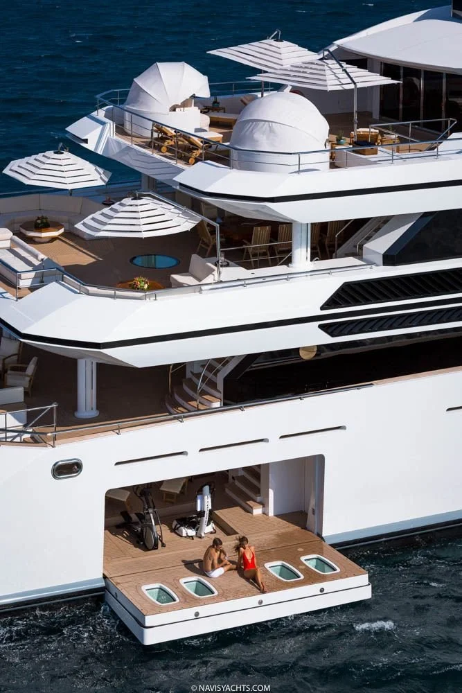 A large white yacht with multiple decks and outdoor seating areas, including sun umbrellas, a hot tub, and two women sitting on the lower deck near the water.