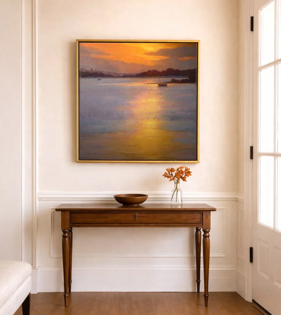 Oil painting of a glowing coastal sunset with a golden shimmer path reflecting across calm water, distant shoreline and boats silhouetted against a warm orange sky, displayed in a refined interior setting above a wooden console table.