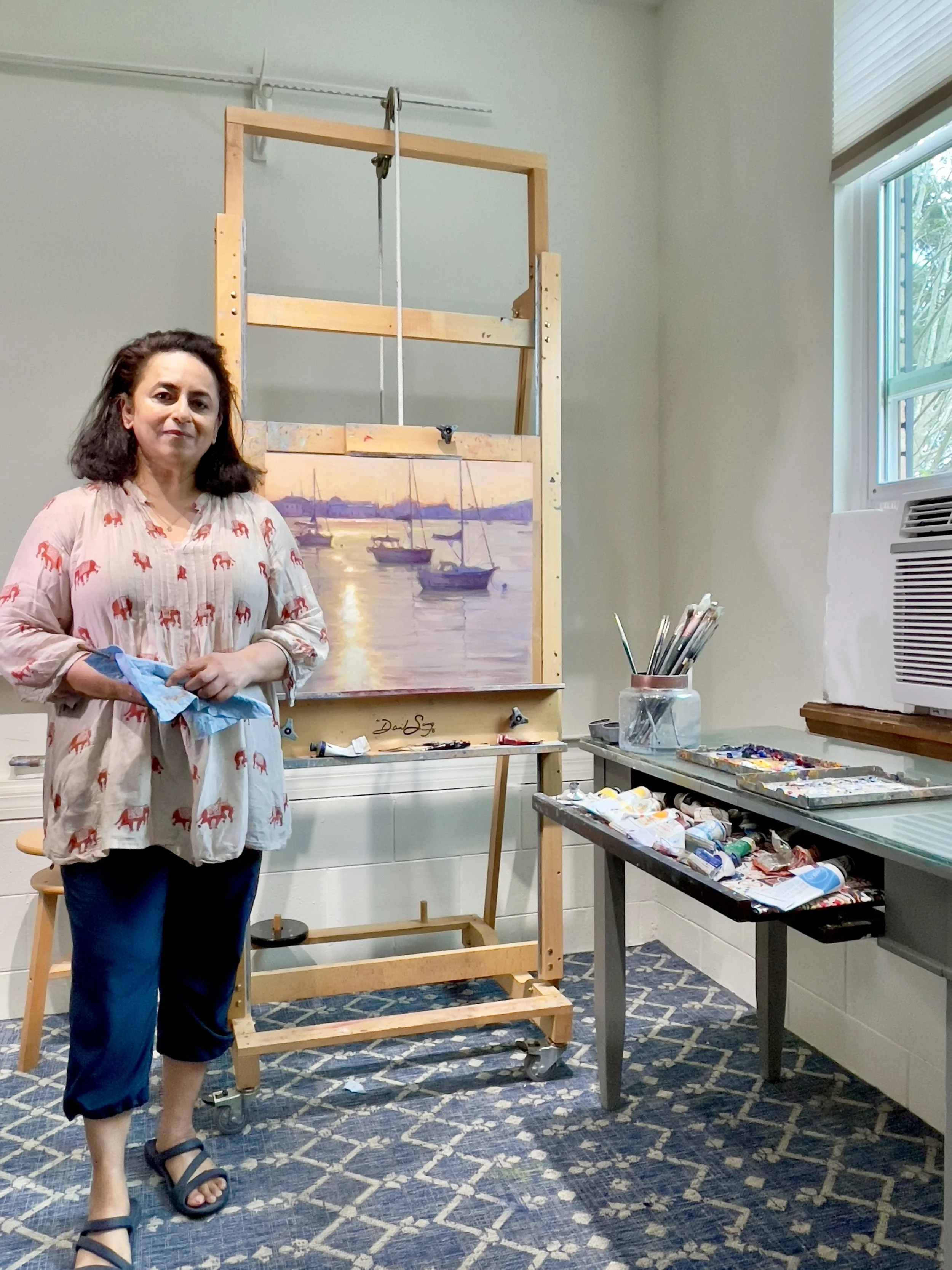 A woman standing in an art studio next to an easel displaying a painting of sailboats on water at sunset. She is wearing a beige blouse with red elephants, navy capris, and black sandals, and is holding a blue cloth. There are art supplies on a table to her right and a small window on the right side of the room.
