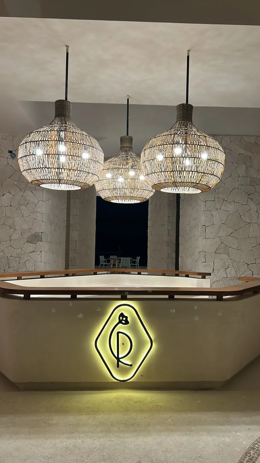Lámparas de mimbre colgantes en el techo y una pared de piedra. Mostrador con un logotipo iluminado en forma de flor o planta.
