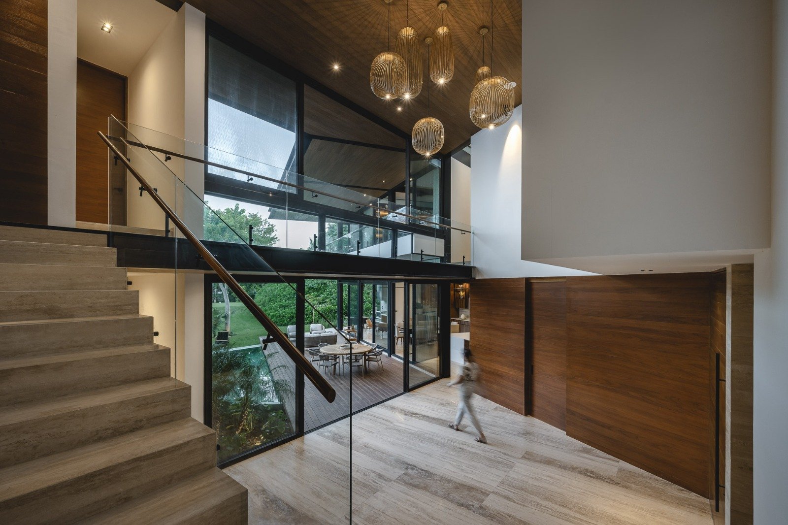 Interior moderno de una casa con escaleras de piedra, paredes de color madera, área de descanso con muebles y ventanas grandes que dejan ver un patio con césped.