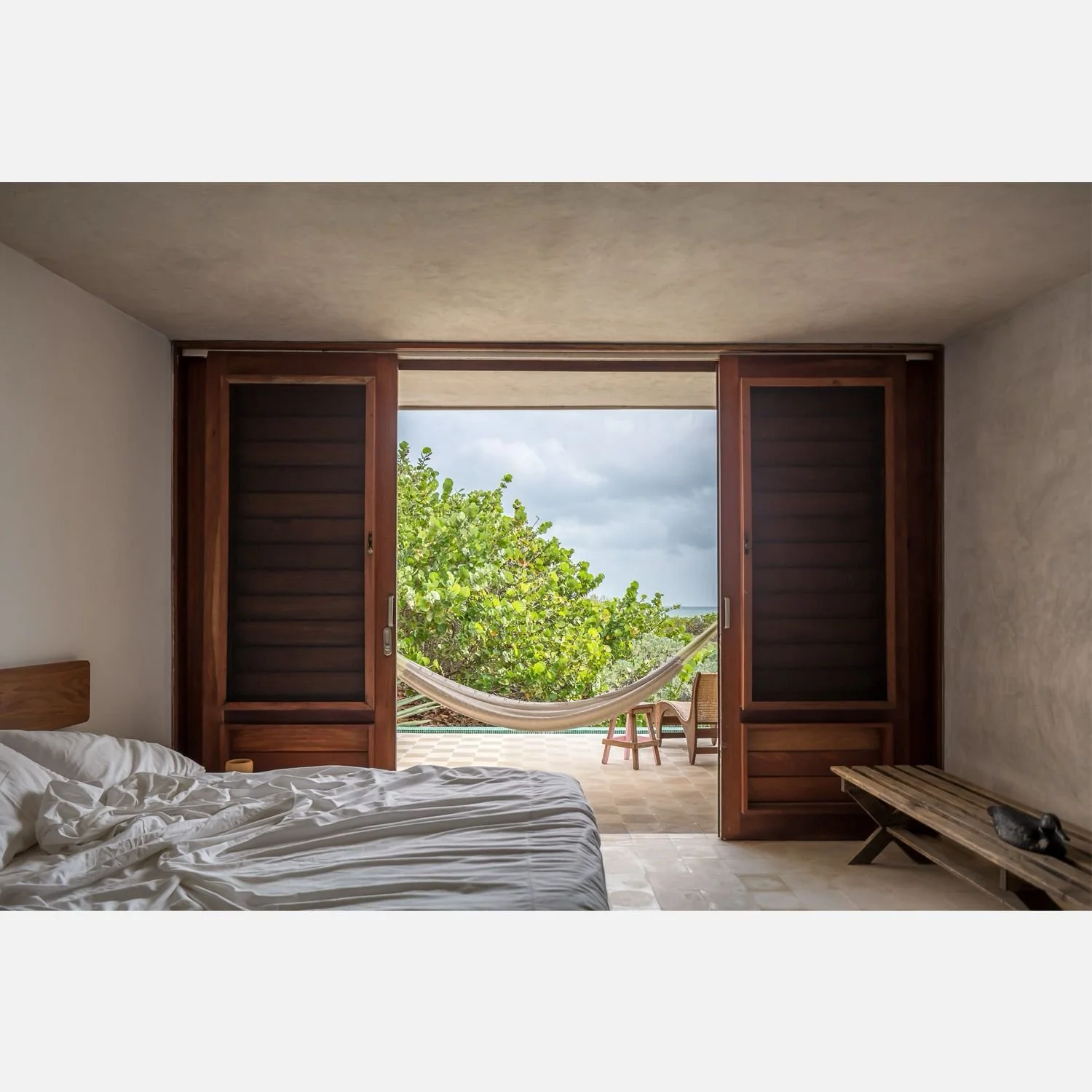 Habitación con cama desordenada, puertas correderas abiertas hacia una terraza con hamaca, vista de árboles y cielo nublado.