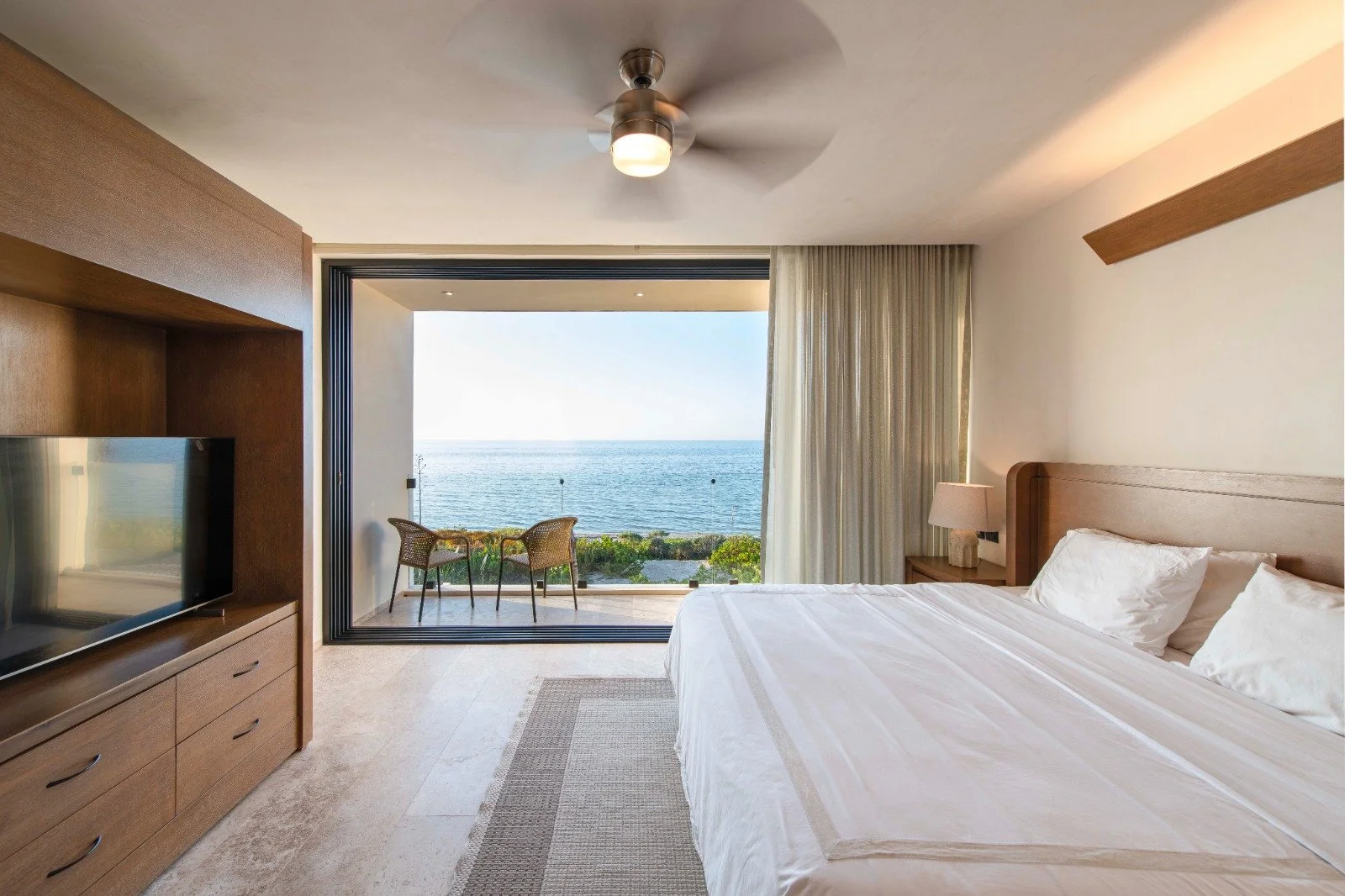 Habitación con vista al mar, cama grande con sábanas blancas, televisor en mueble de madera, balcón con sillones y vista al océano, cortinas claras.