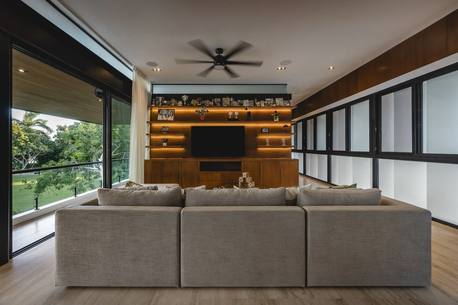 Sala moderna con sillón beige, mueble de madera con pantalla y estanterías iluminadas, paredes de vidrio con vista a árboles verdes.