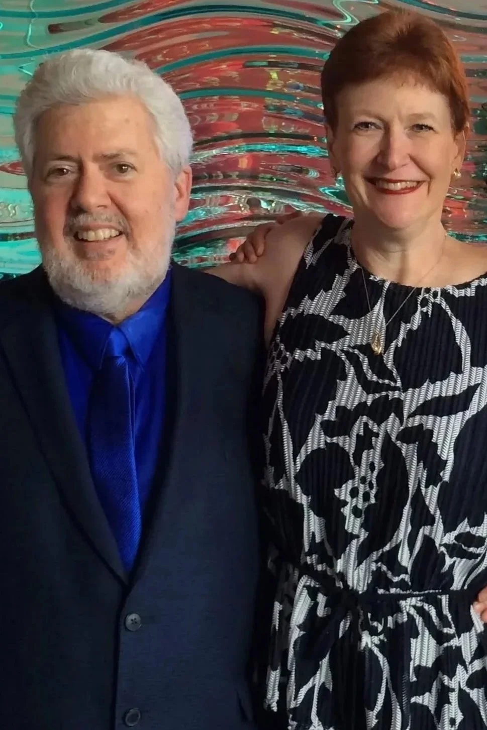 A smiling man with a white beard in a black suit and blue shirt, and a smiling woman with short red hair in a sleeveless black and white patterned dress, standing together in front of a colorful, abstract, swirling background.