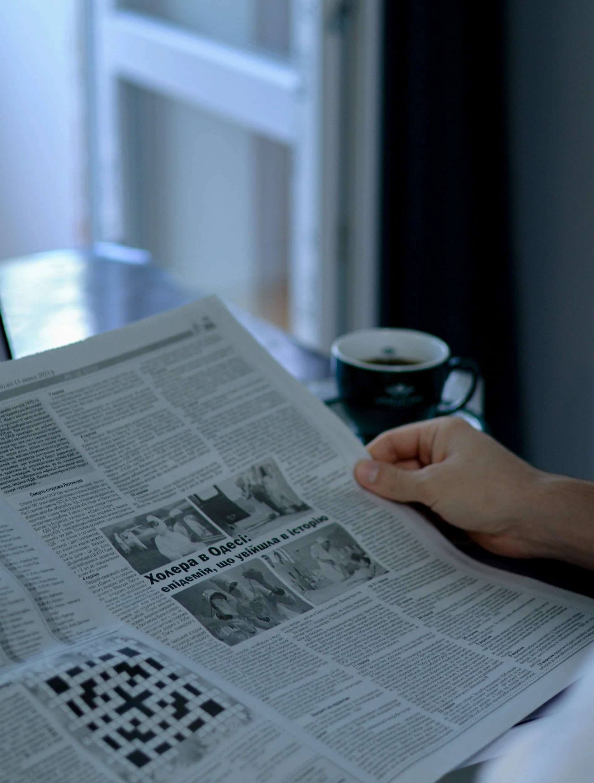 Person holding a newspaper with a black cup of coffee in the background, near a window.