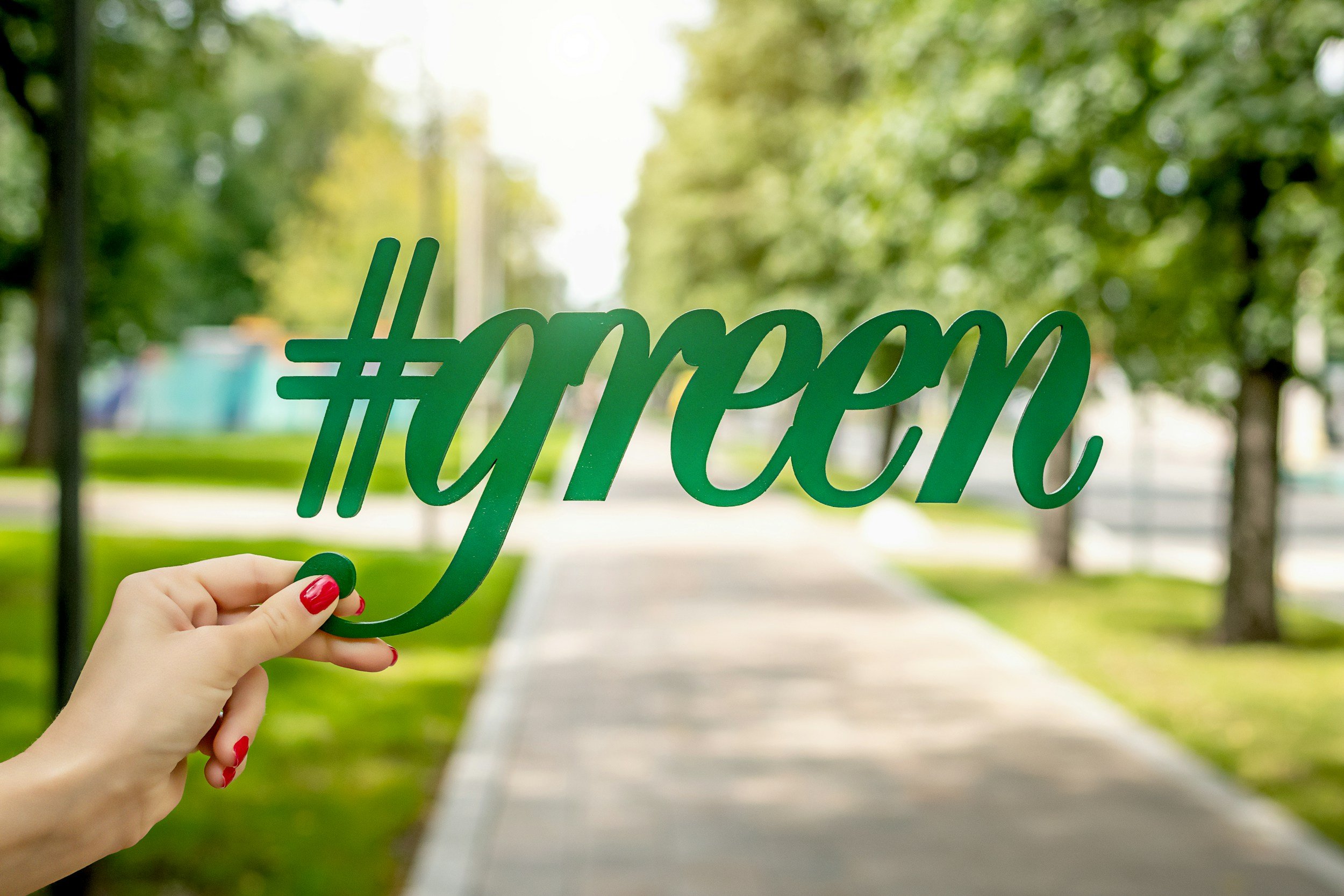 A person's hand with red painted nails holding a green hashtag sign that reads '#payen' on a sidewalk in a park-like area with green trees in the background.