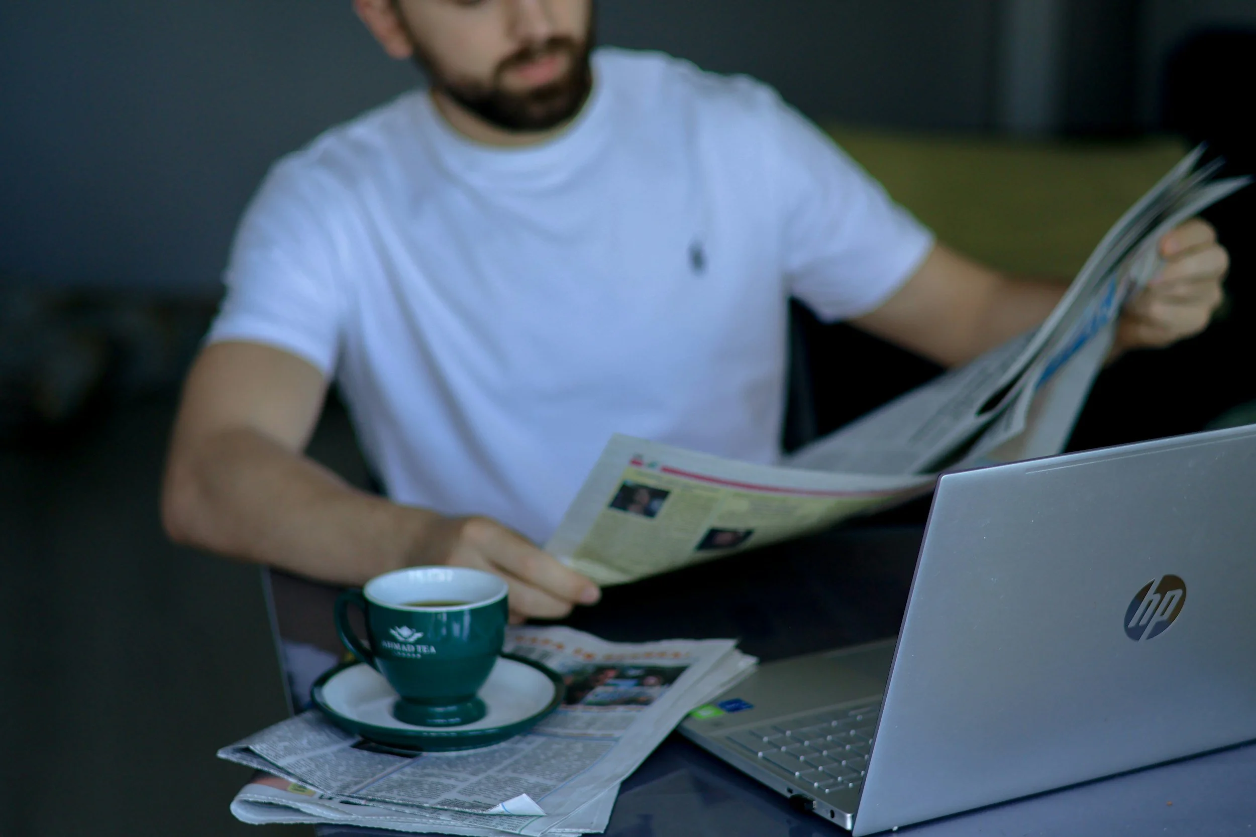 Man in a white t-shirt reading a newspaper at a black table, with a silver HP laptop, a cup of coffee, and newspapers spread out.
