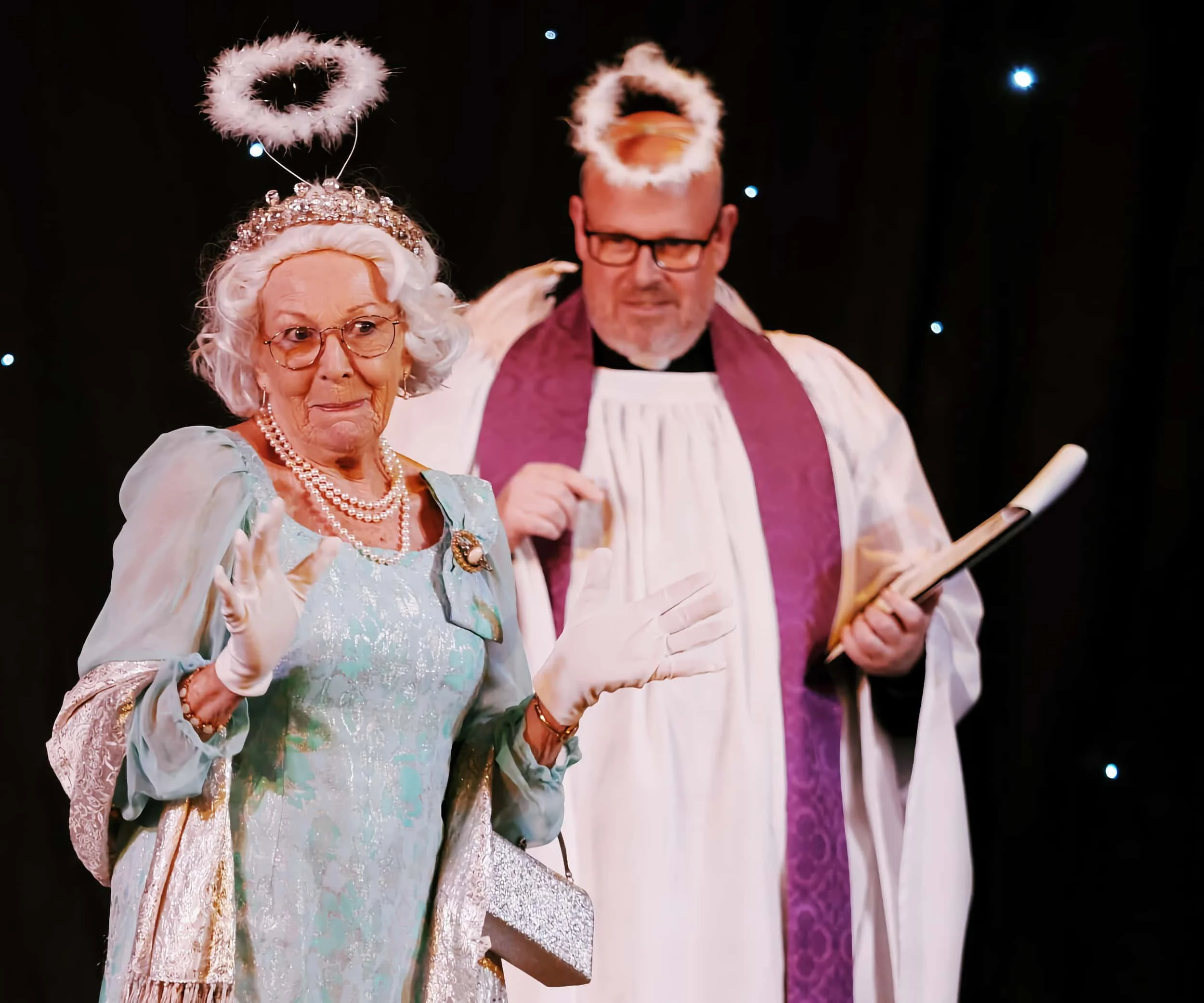 An elderly woman dressed as an angel with a halo, tiara, pearl necklace, glasses, and a fancy dress, standing next to a man dressed as a priest with a halo, robes, and holding a book, against a dark background with small lights.