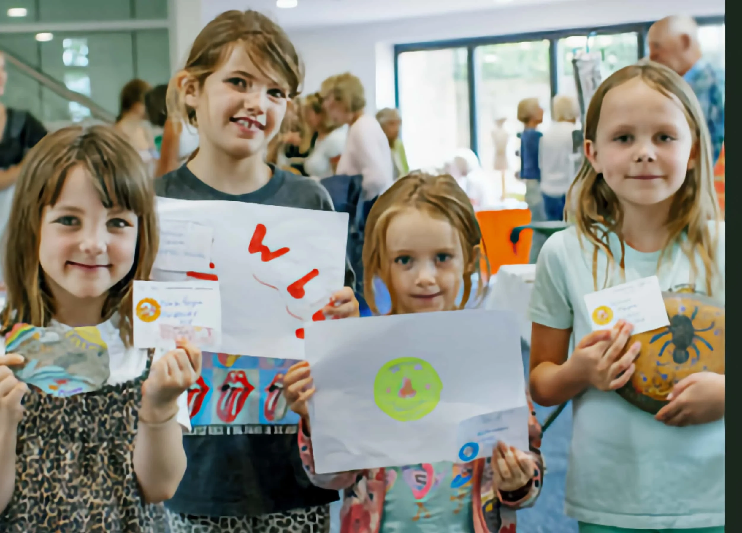 Four young girls holding colorful drawings and art projects in a bright indoor space, with other people and art displays visible in the background.