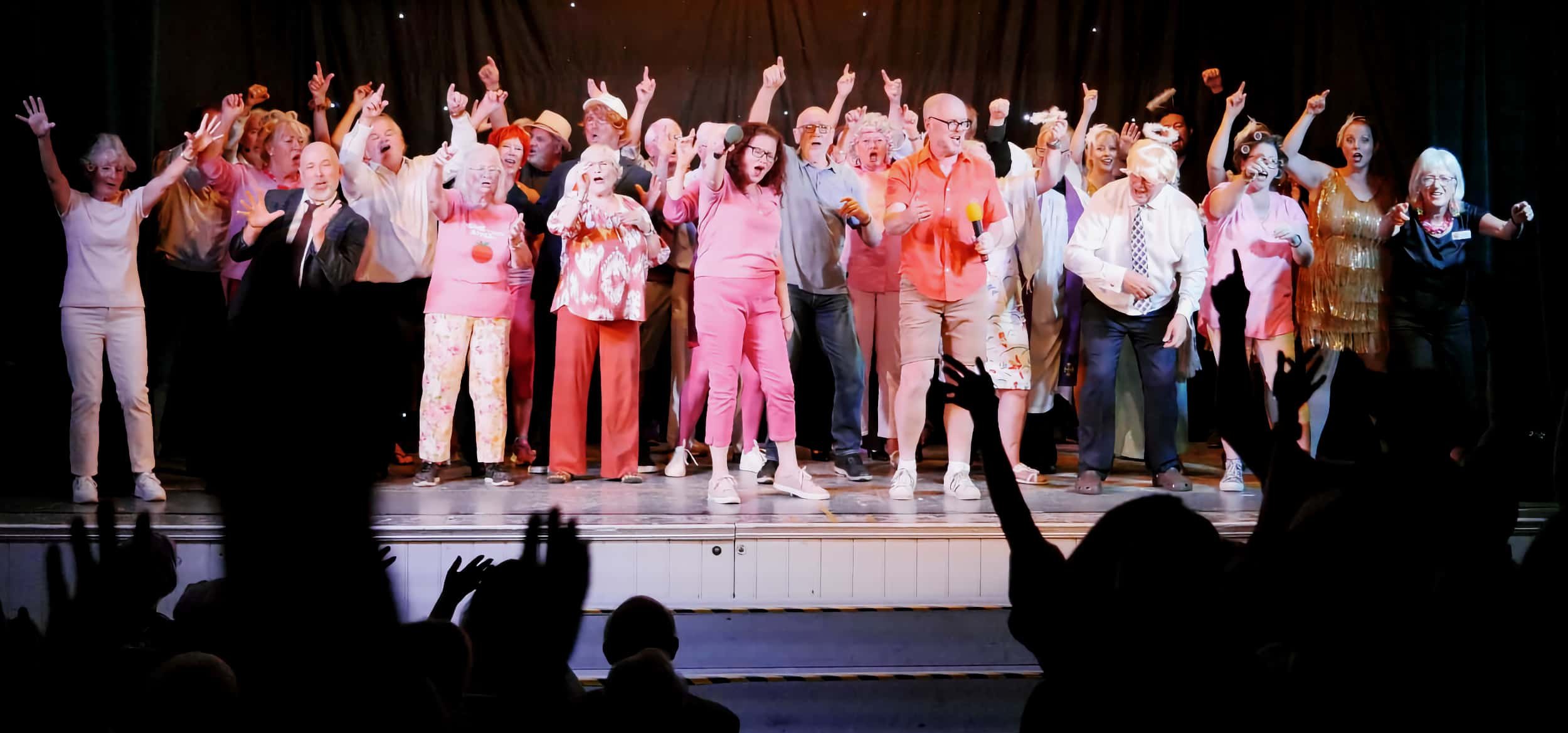 A large group of elderly people on stage singing and dancing, with an audience clapping and cheering in the foreground.
