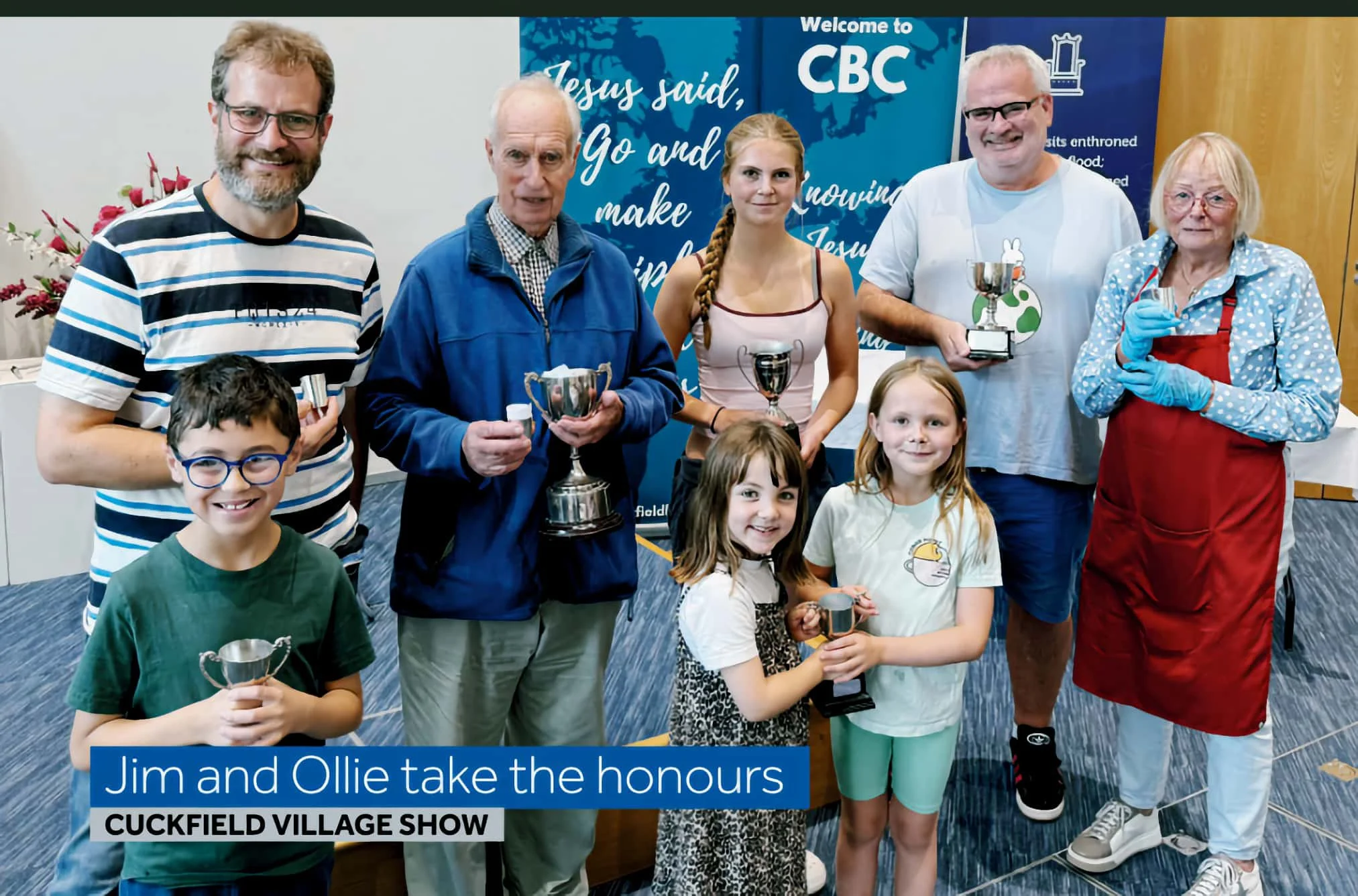Group of children and adults at an awards ceremony, holding trophies, with banners in the background that read 'Welcome to CBC' and 'Jesus said, Go and make disciples.' The caption states 'Jim and Ollie take the honours at Cuckfield Village Show'.