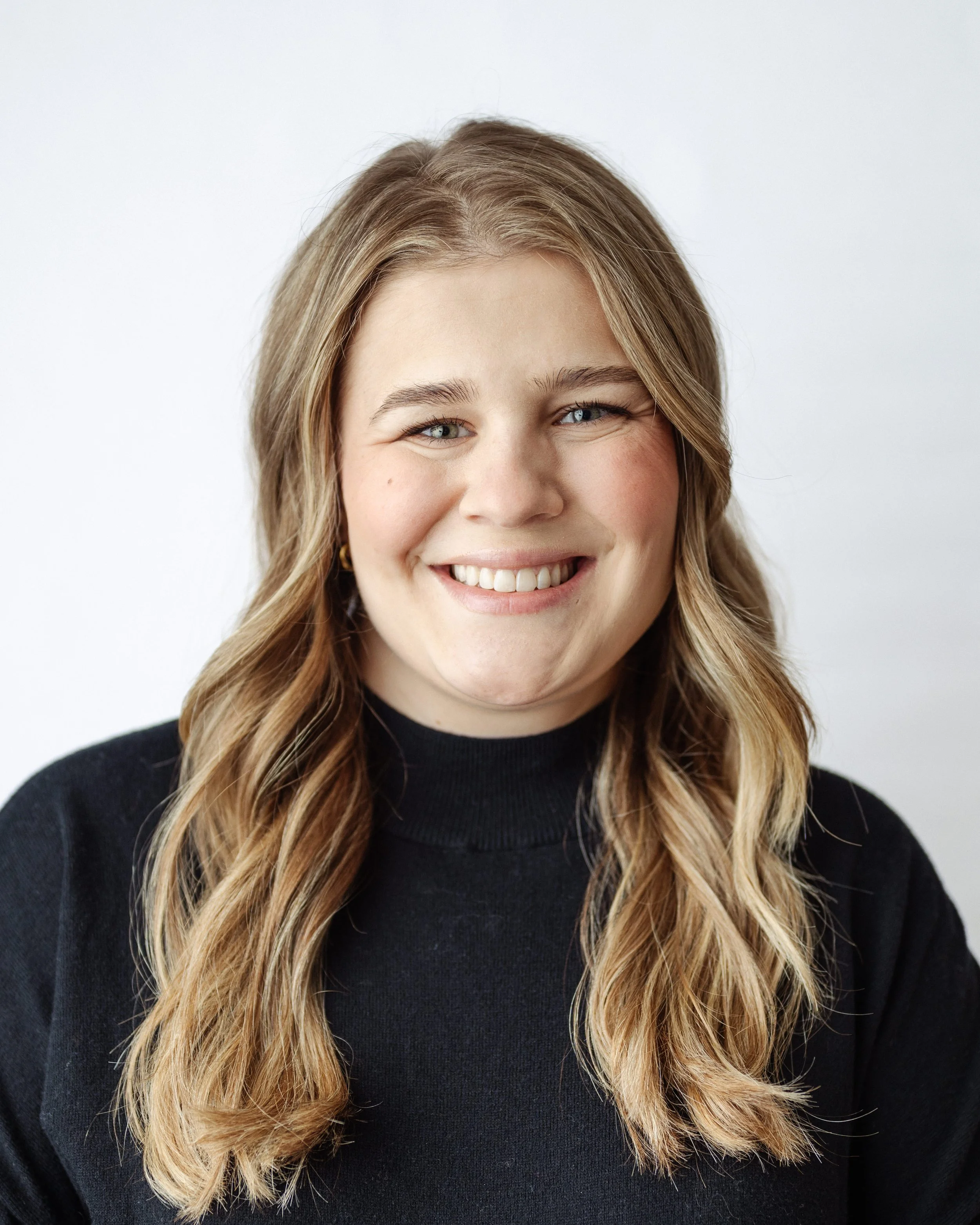 Portrait of a young female therapist. Providing therapy to those looking for help with trauma, life transitions also addiction. Therapist with long wavy blonde hair, wearing a black turtleneck sweater, smiling against a plain white background.