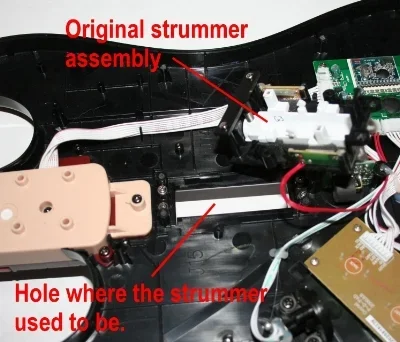 Close-up photo of an electronic device's interior showing an original strummer assembly and the hole where the strummer used to be, with red annotations highlighting these parts.