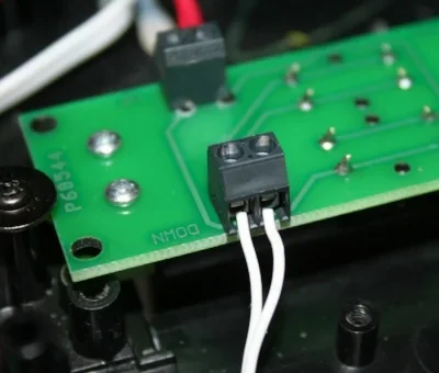 Close-up of a green printed circuit board with electrical components and two white wires connected to a black connector.