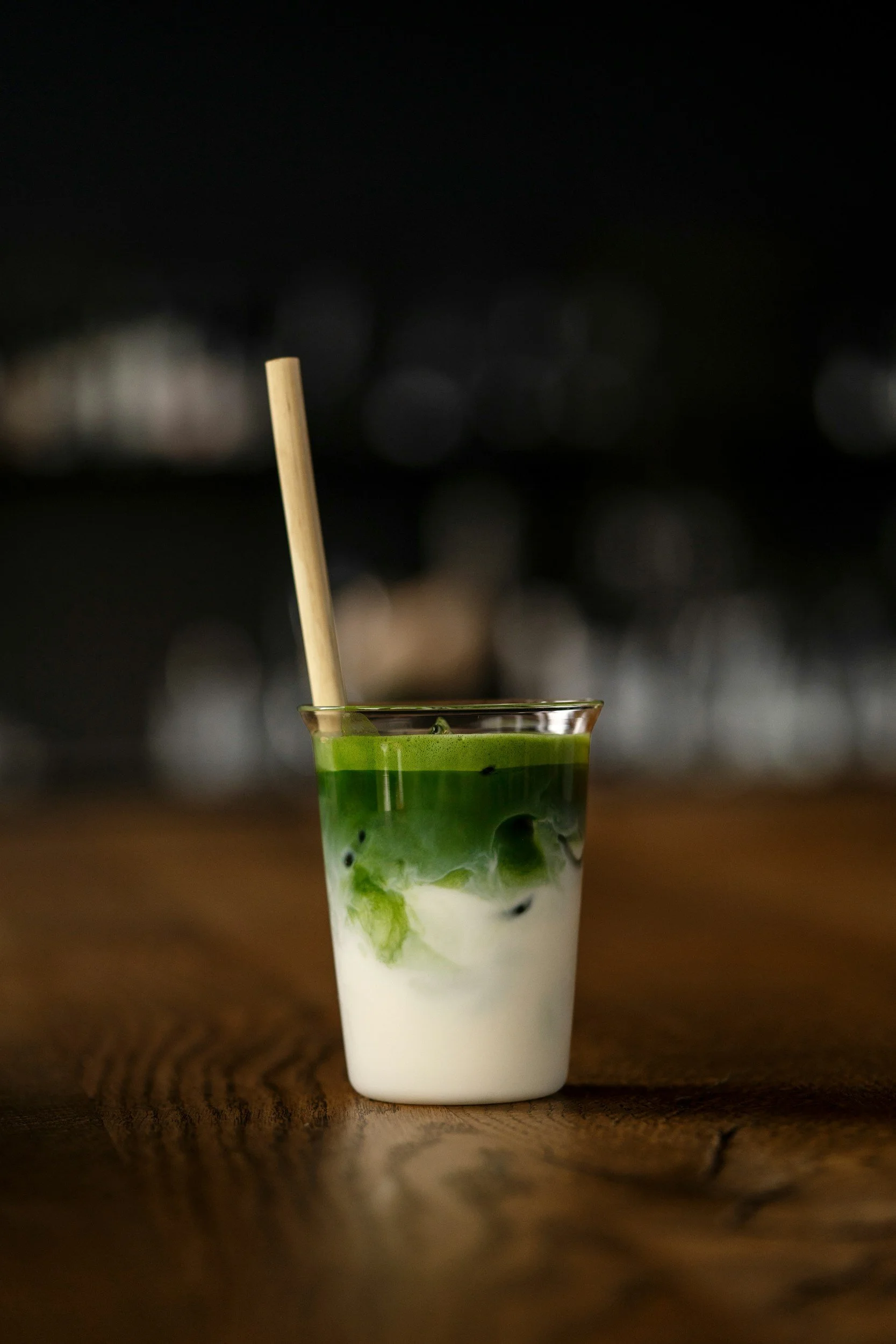 A clear glass containing a layered drink with white milk at the bottom and bright green matcha tea or powder on top, with ice cubes and a bamboo straw. Coffee catering event in Omaha, NE by Ahamo Coffee Co.