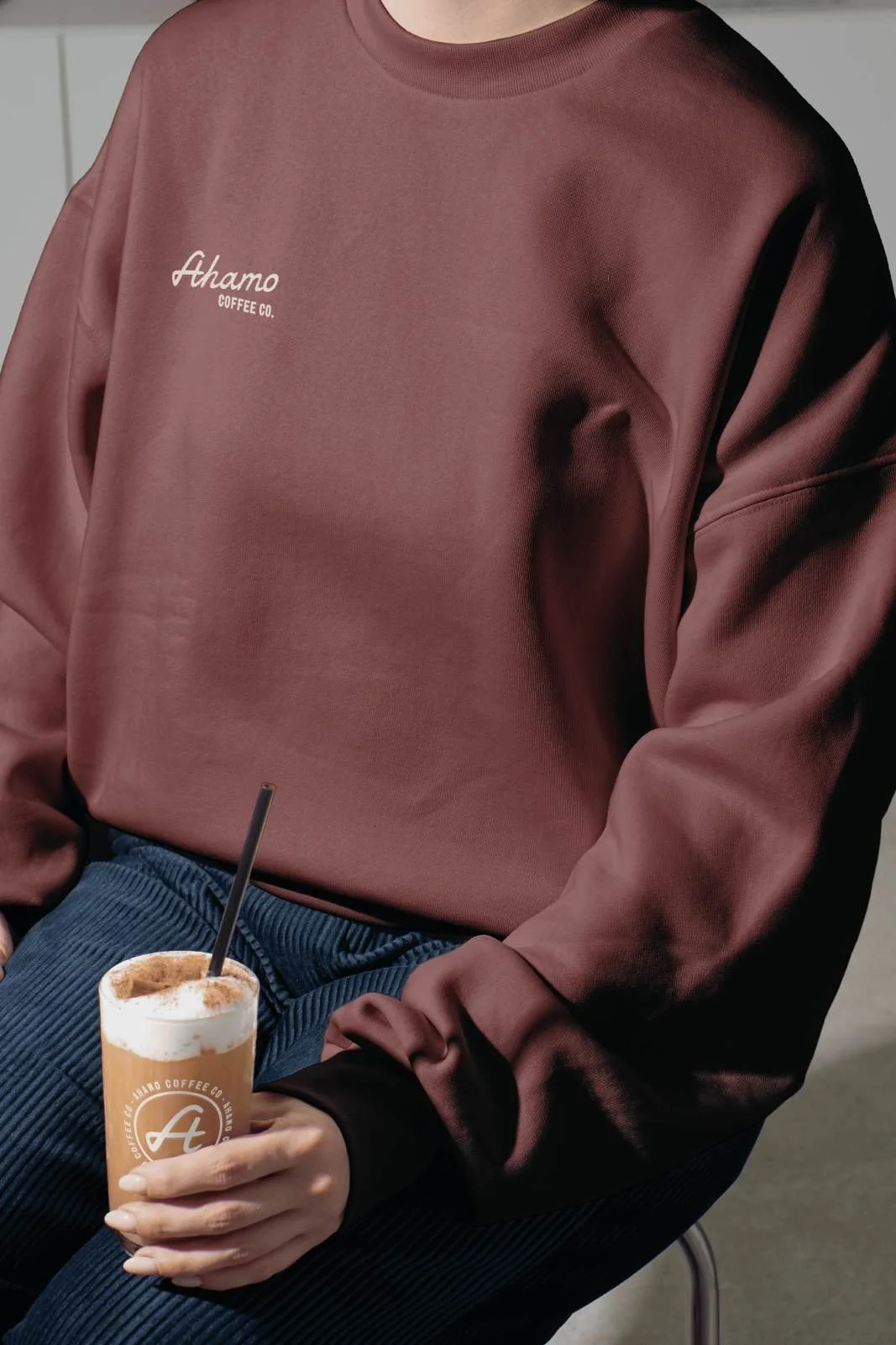 Person in a mauve sweatshirt with 'Ahamo Coffee Co.' logo sitting with legs crossed, holding a glass of iced coffee at an event in Omaha, NE.