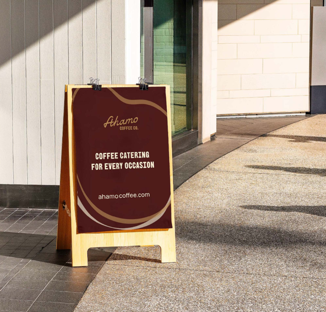 A wooden A-frame sidewalk sign with a maroon background and white text advertising Ahamo Coffee Co. and their coffee catering services outside a building with glass doors in Omaha, NE.