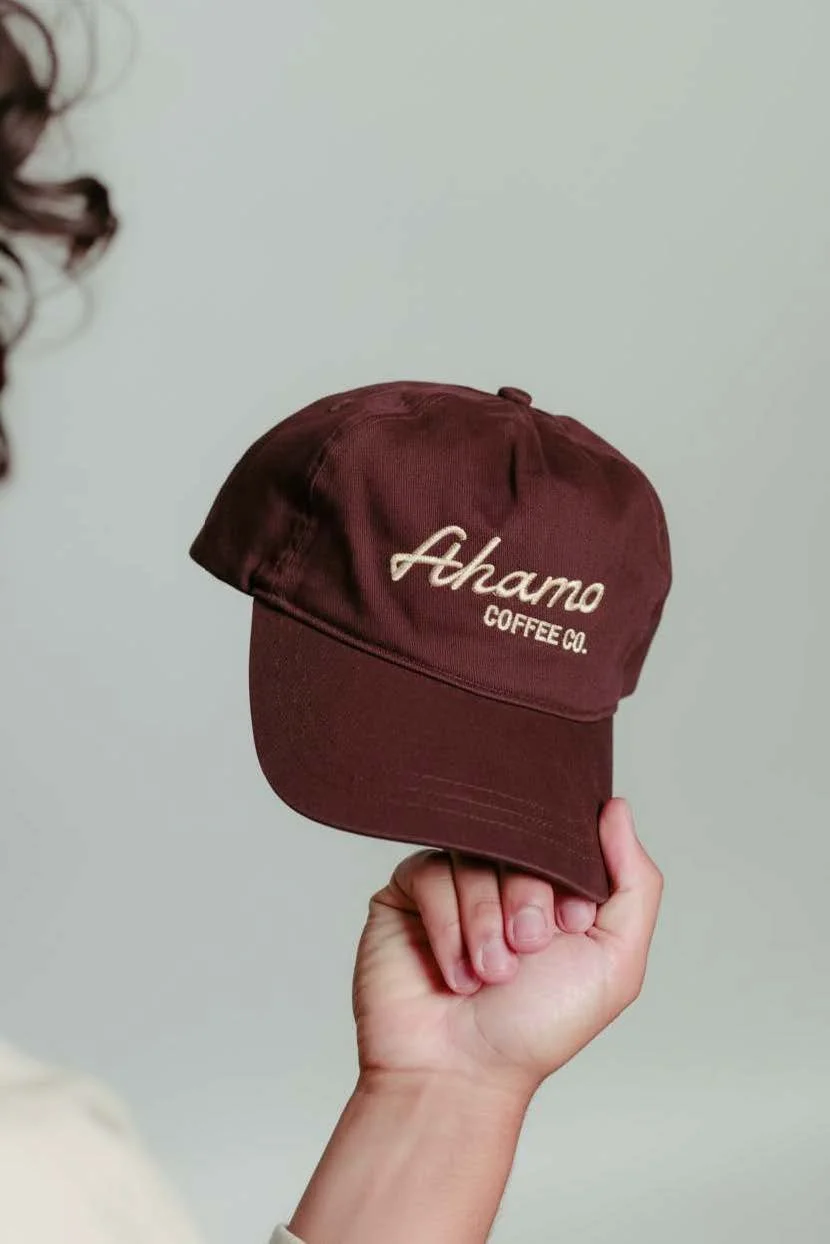 A person holding a maroon baseball cap with embroidered white text that reads 'Ahamo Coffee Co.', a coffee catering service in Omaha, NE.