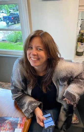 Woman with brown hair smiling and holding a smartphone, sitting at a kitchen table near a window with a view of a driveway and parked cars.