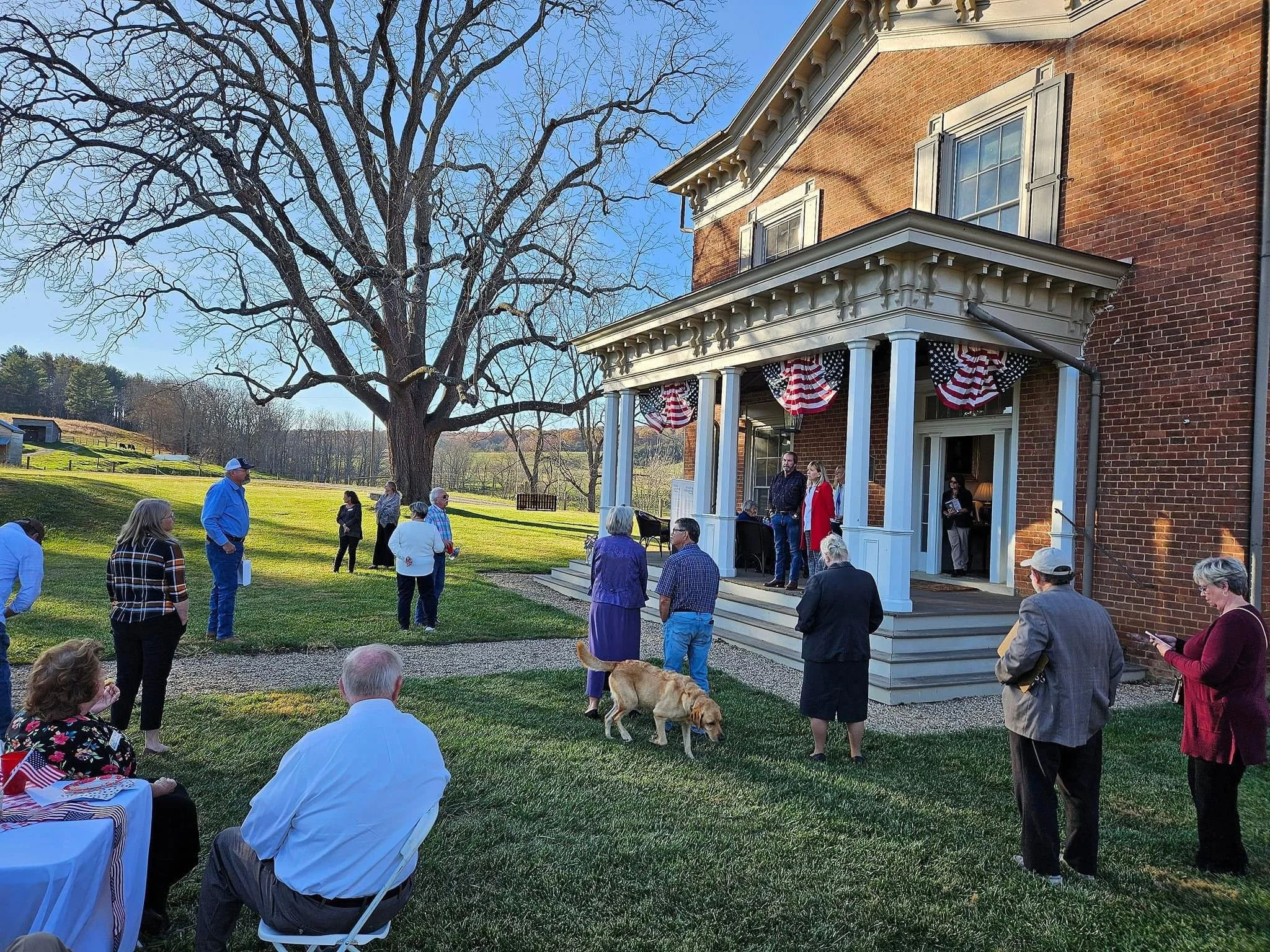 People gathered on a lawn and front porch for an outdoor event at a brick house decorated with American flags and patriotic bunting. Some people are sitting, some are standing and talking, and a dog is present. The scene is set in the late afternoon with a large leafless tree and rural landscape in the background.