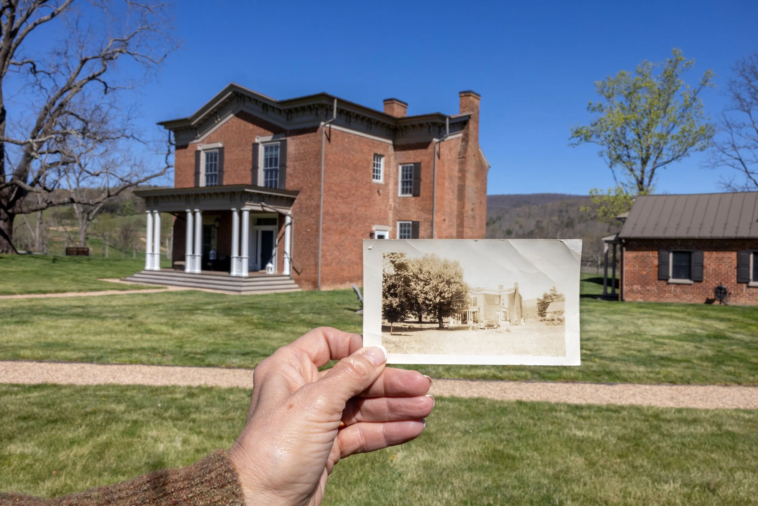 Person holding an old black-and-white photograph of a house in front of a modern brick house with a porch, set on a grassy area with trees.