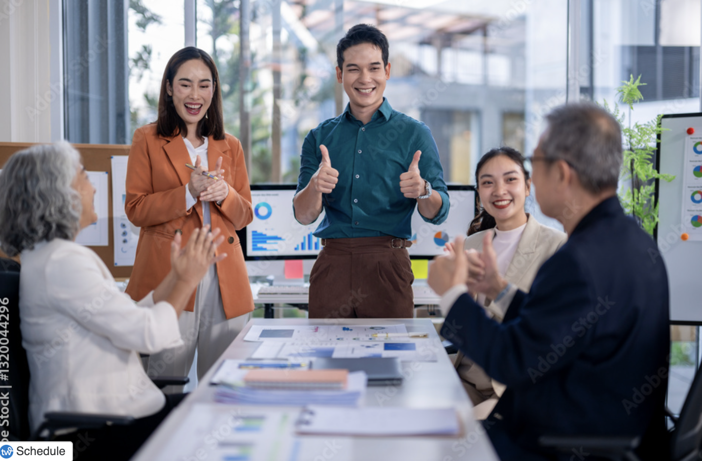 A diverse group of five business professionals in a modern office, smiling and conversing, with two standing and three sitting at a conference table with charts and documents, during a meeting.
