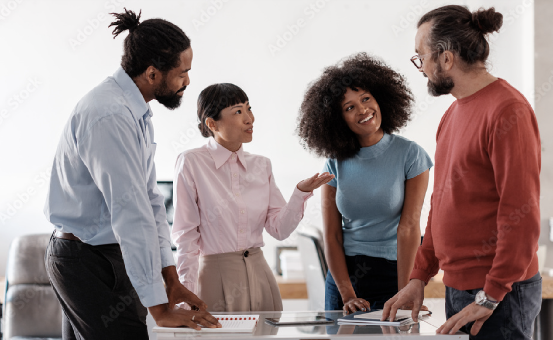 Five professionals in a meeting room engaged in discussion, standing around a table with laptops and documents, with positive expressions and gestures.
