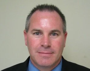 Close-up of a man with short, dark hair, wearing a dark suit and blue shirt, smiling slightly against a plain, light-colored background.
