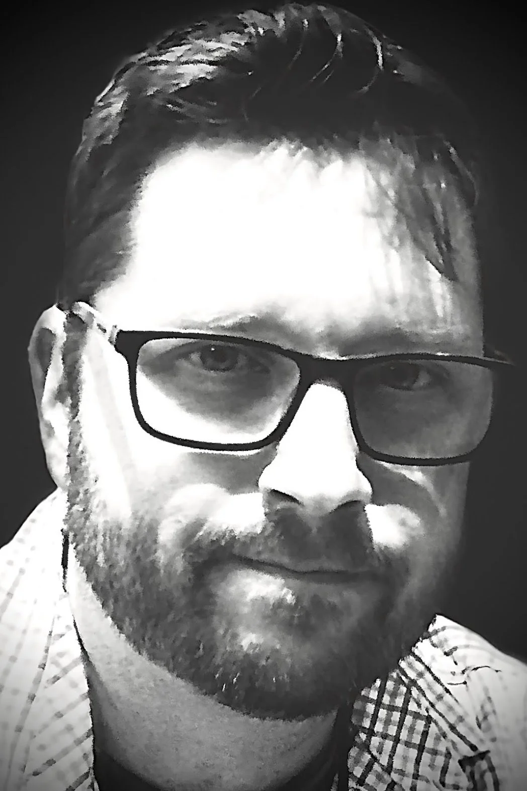 A black and white photo of a man with glasses, wearing a checkered shirt, adjusting his glasses with his right hand.