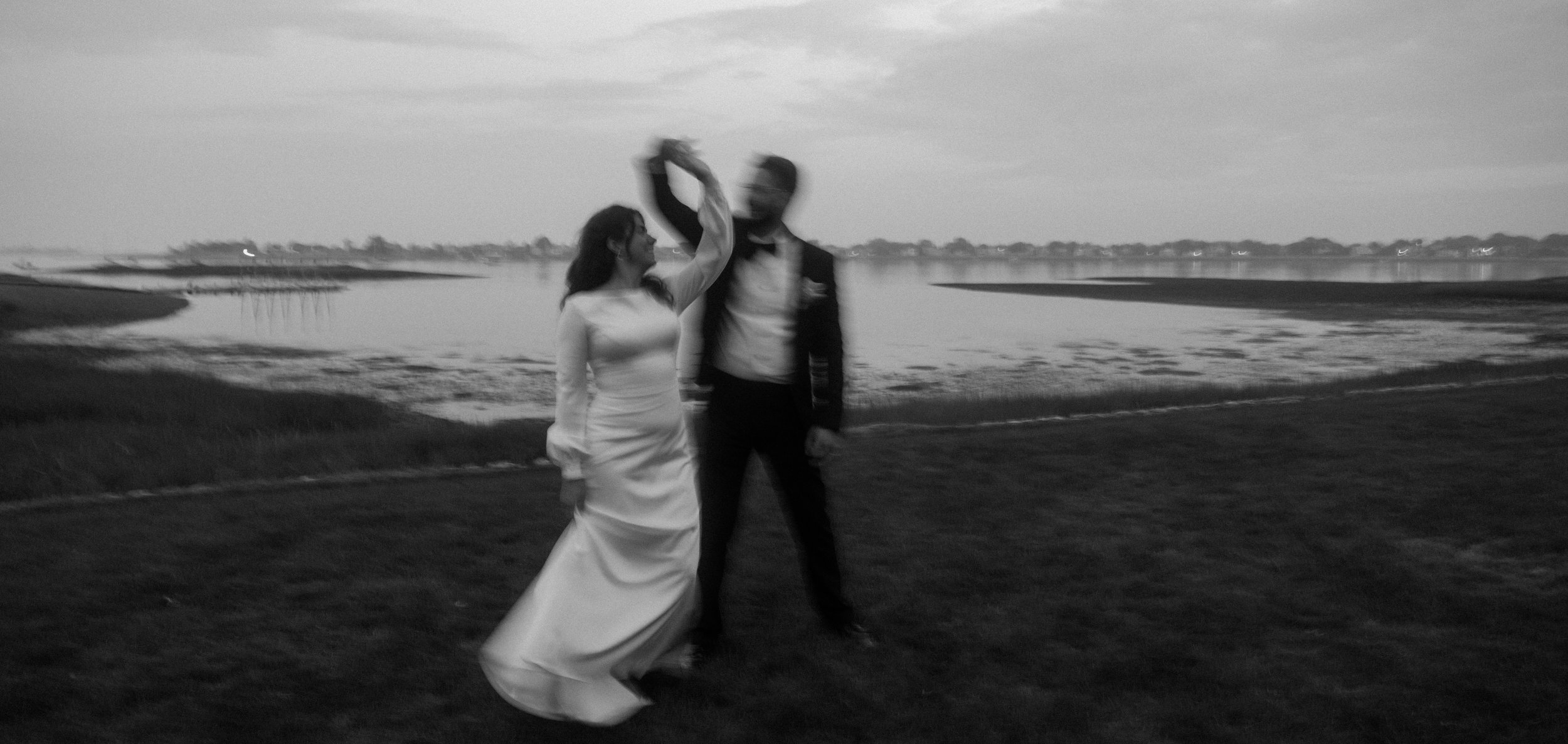 A couple dressed in formal attire dancing by a lakeside at sunset in black and white.
