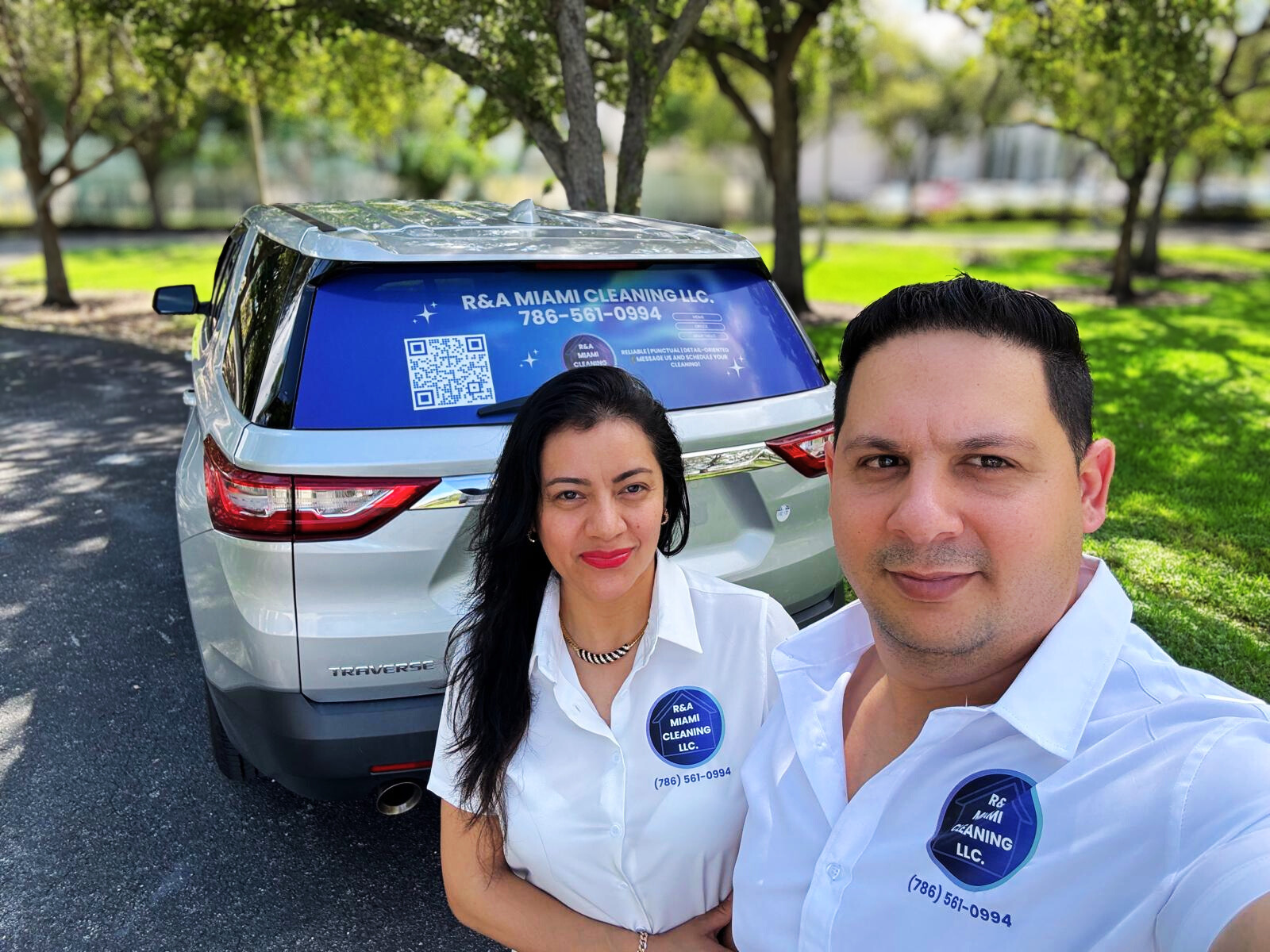 A man and woman taking a selfie outdoors in front of a silver SUV for R&A Miami Cleaning LLC, with company branding on the vehicle and their shirts, in a park with green grass and trees.