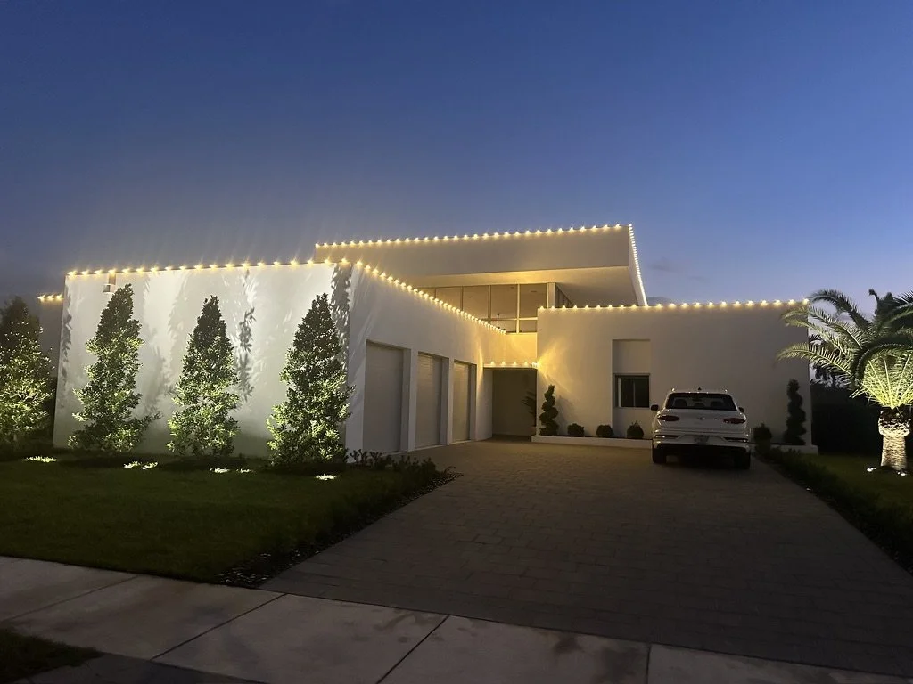 Modern house with white exterior, lit with string lights along the roofline, surrounded by landscaped yard with trees and shrubs, parked car in driveway, during evening.
