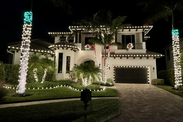 Night view of a two-story house decorated with Christmas lights, including winding lights on the roofline and wrapped around palm trees, with festive wreaths on the upper windows and a paved driveway.