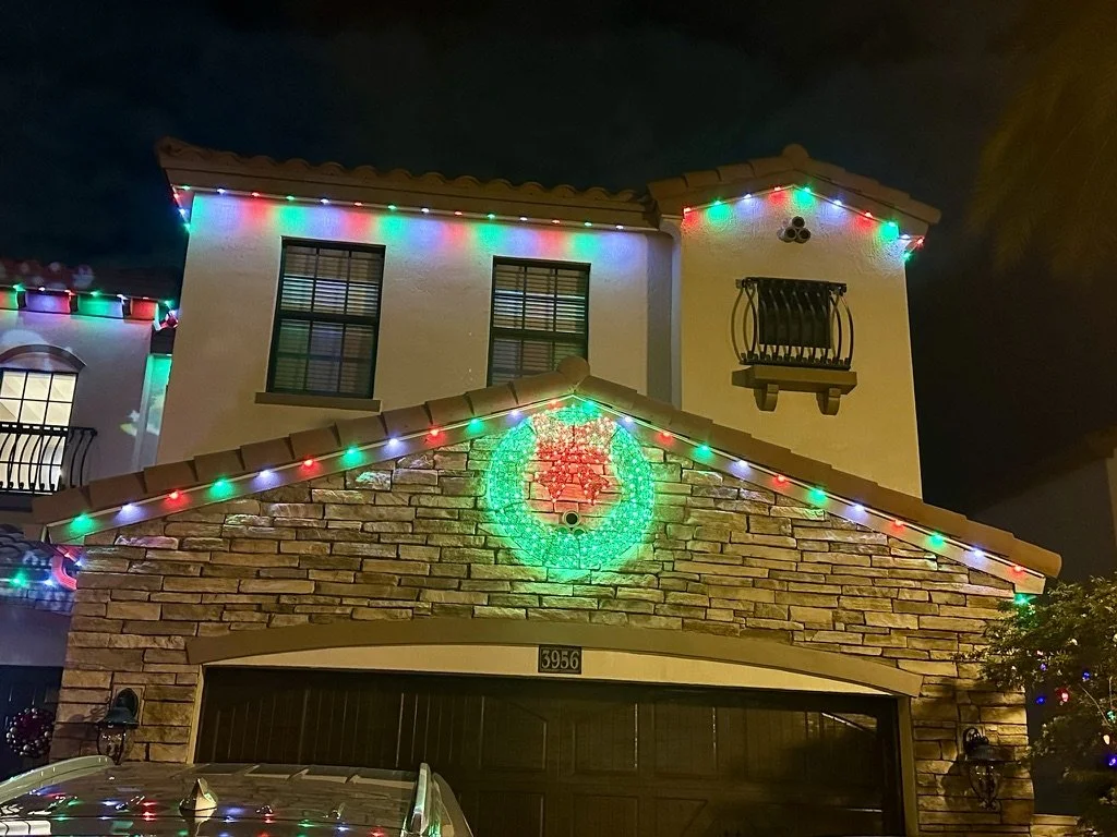 A house decorated with colorful Christmas lights and a green and red wreath design on the brick garage facade at night.
