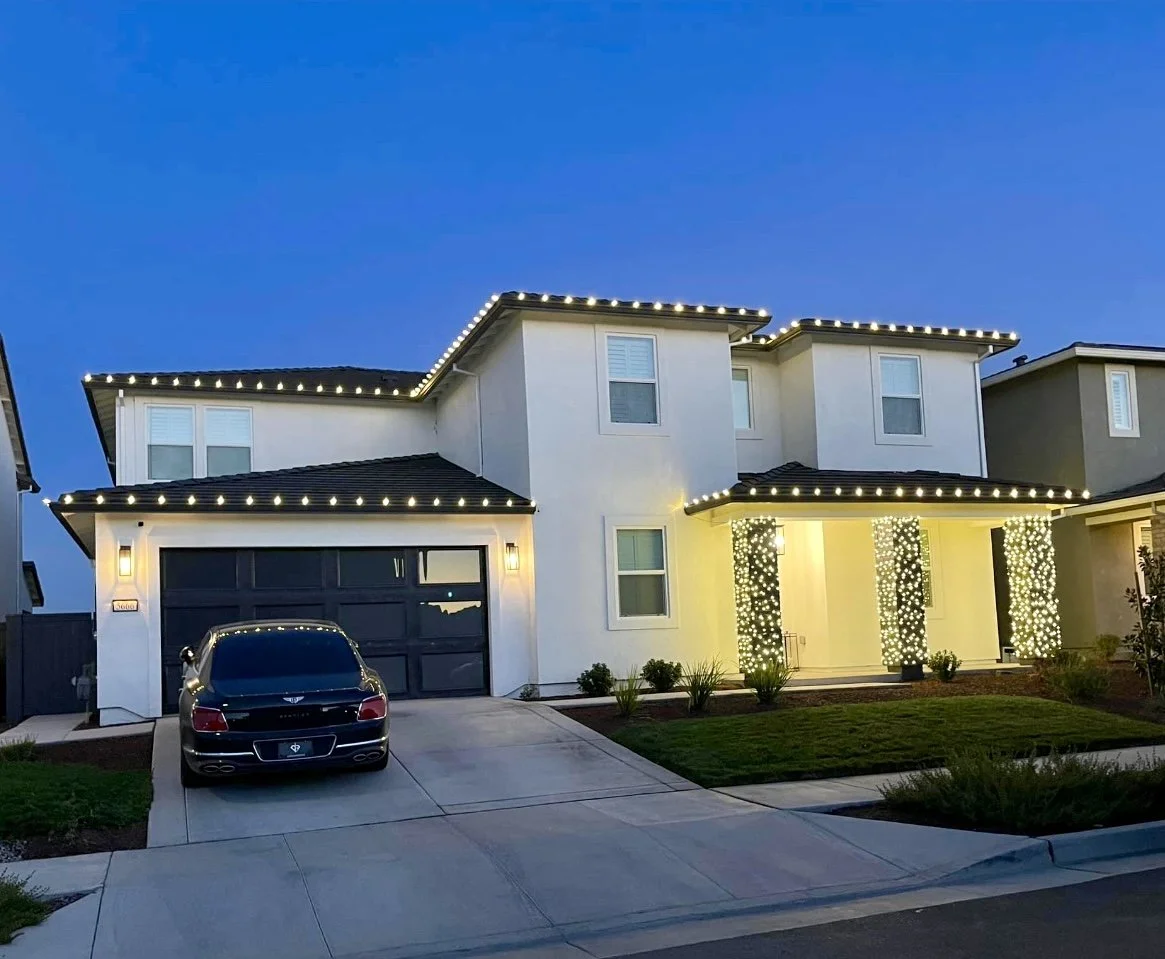 Modern two-story white house decorated with string lights on the roof and pillars, with a black car parked in the driveway during twilight.